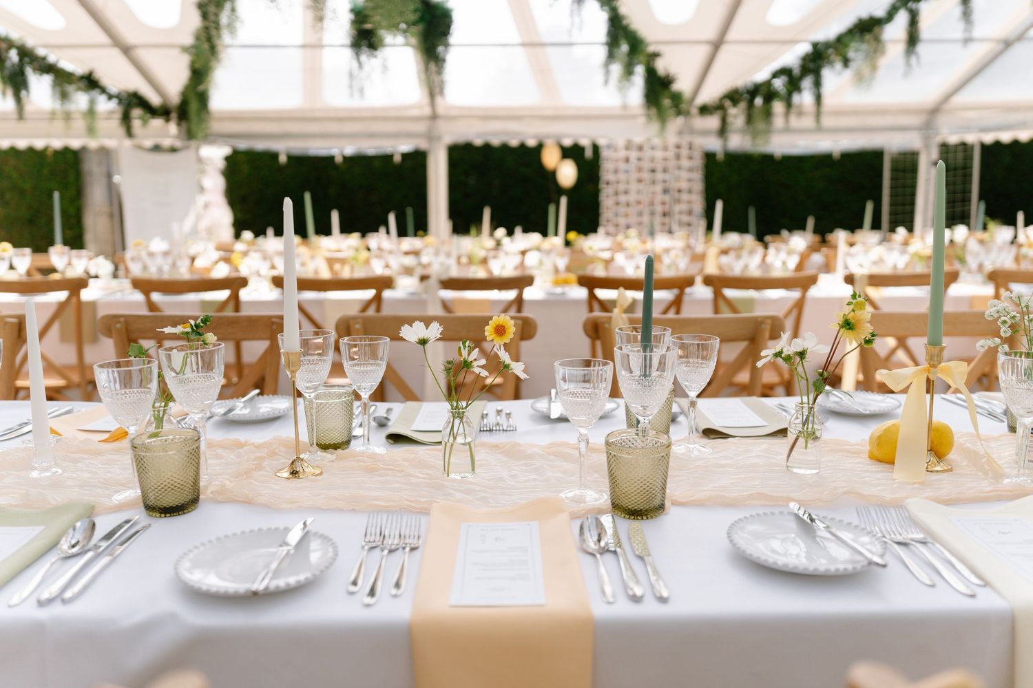Long banquet tables with white linens and place settings under a tent with hanging greenery.