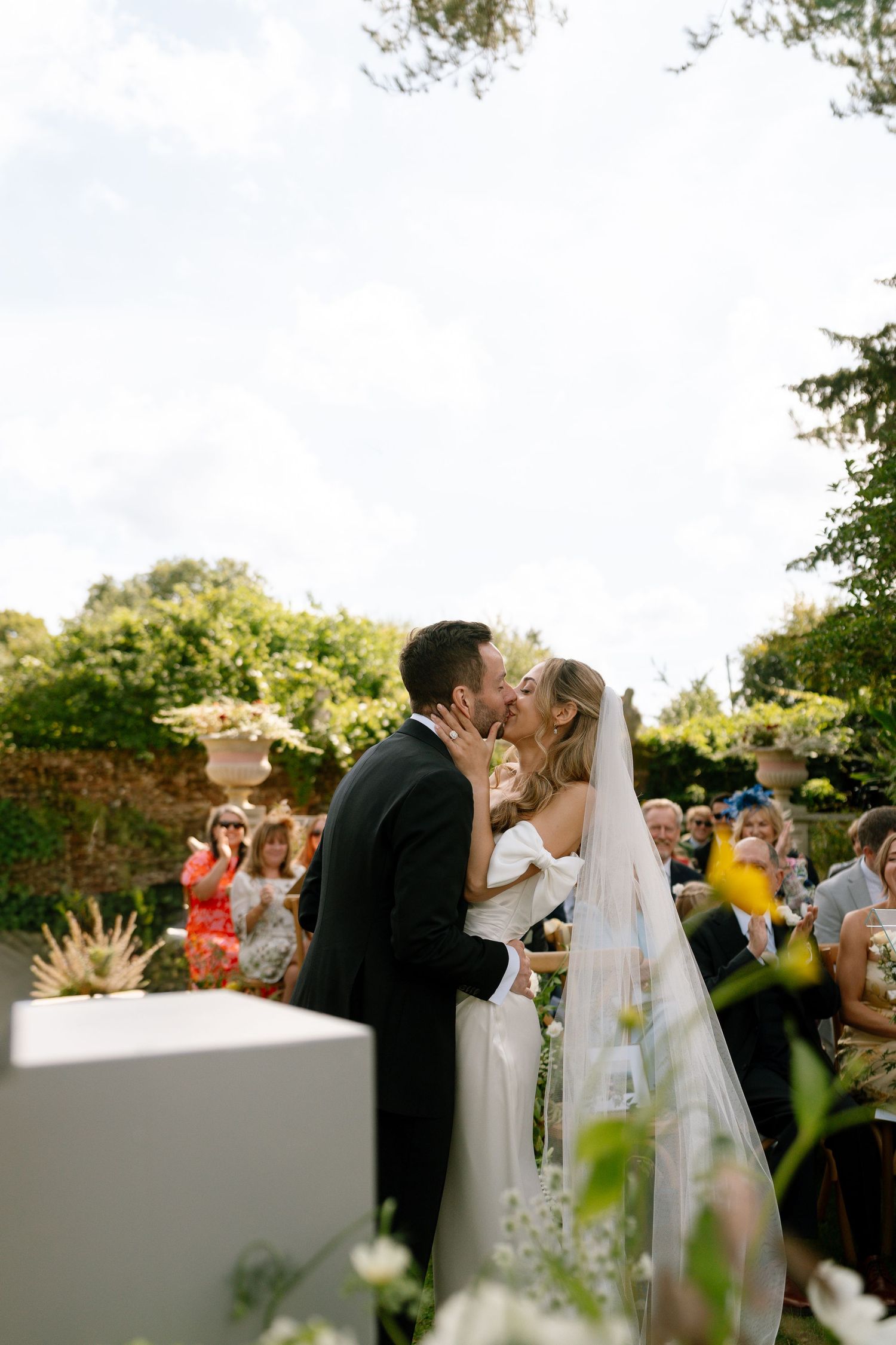 Couple kisses at their outdoor wedding ceremony surrounded by guests and greenery.