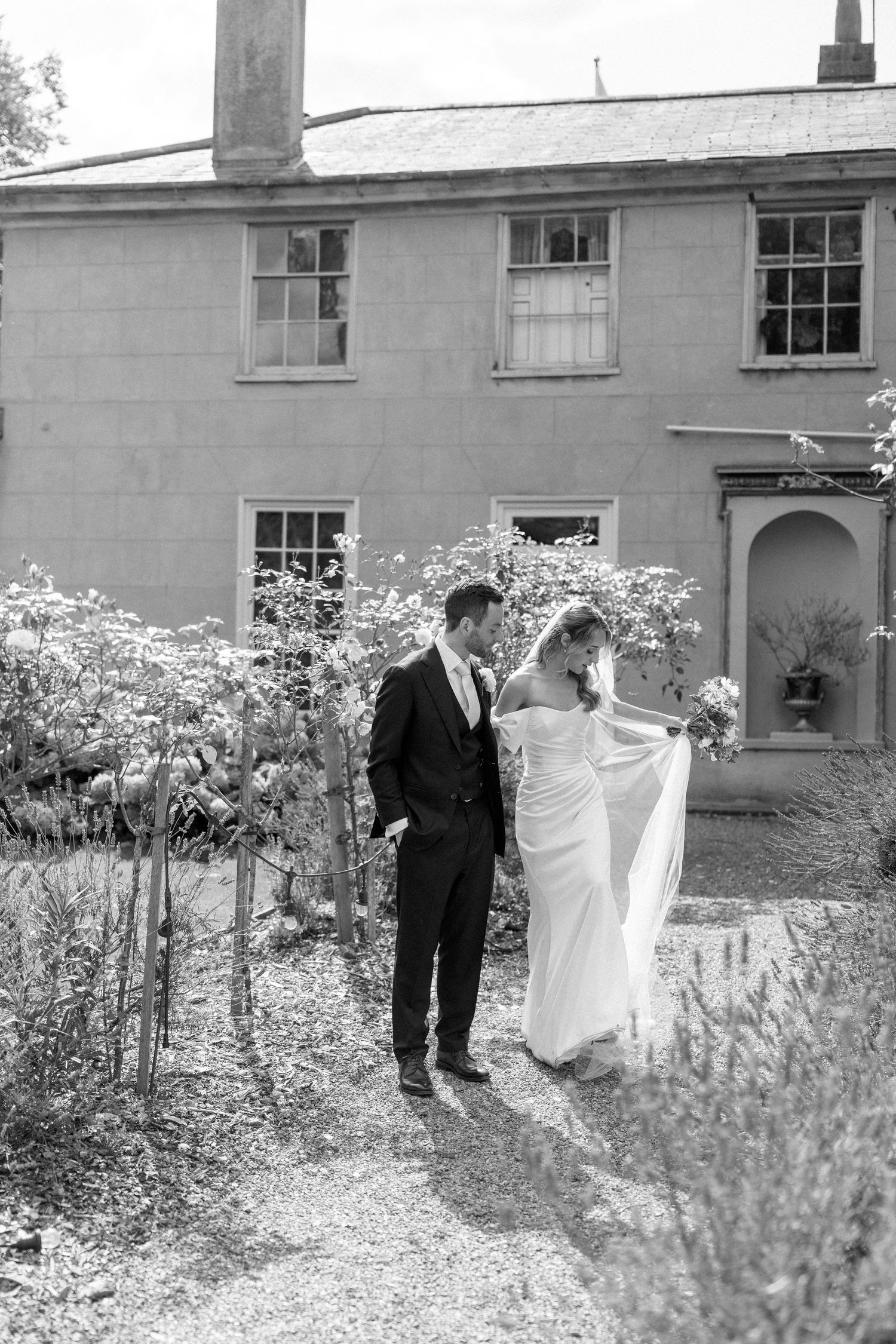 Black and white wedding portraits among flowering garden plants near historic building.