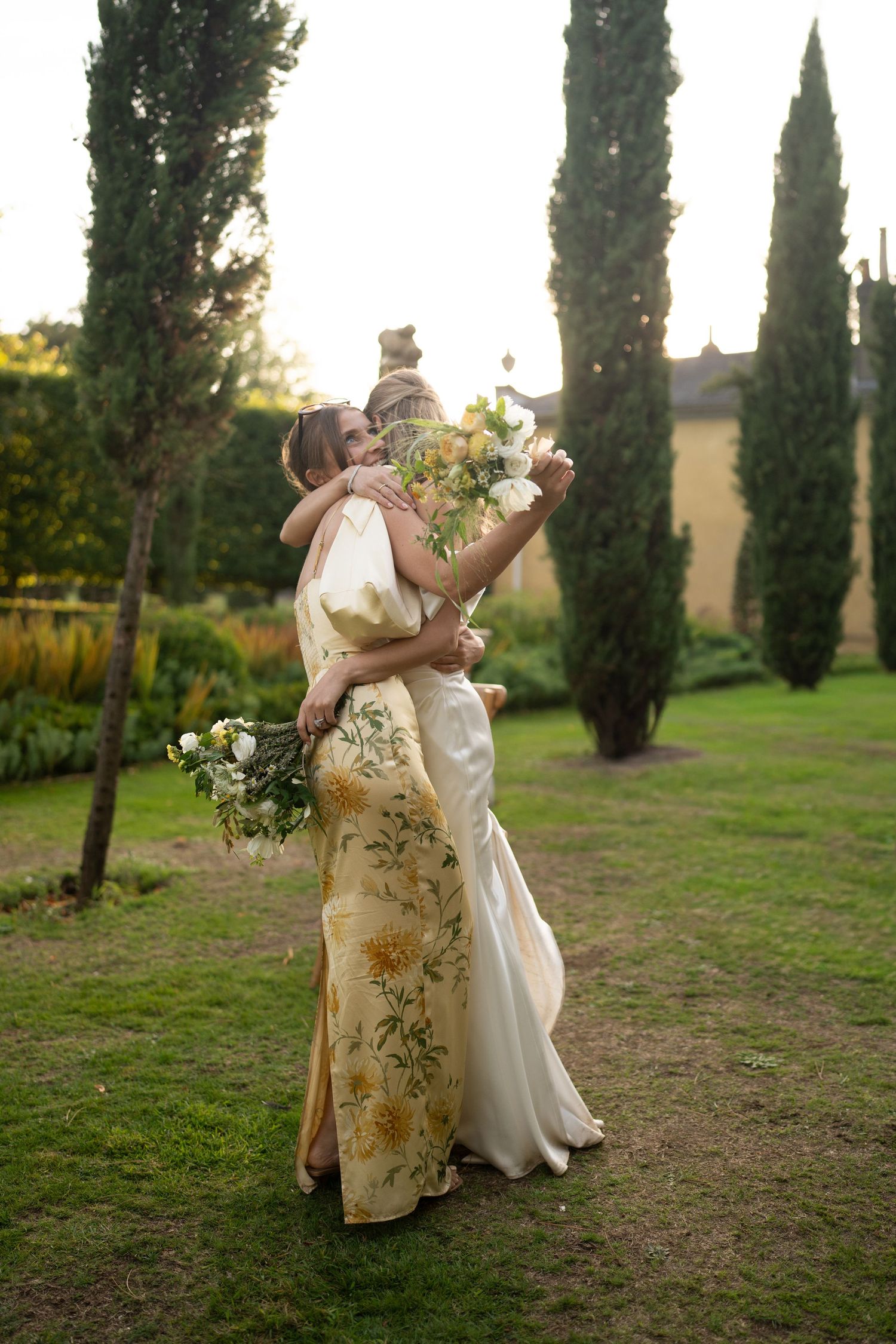 Wedding couple embraces in golden sunlight among cypress trees in garden setting.