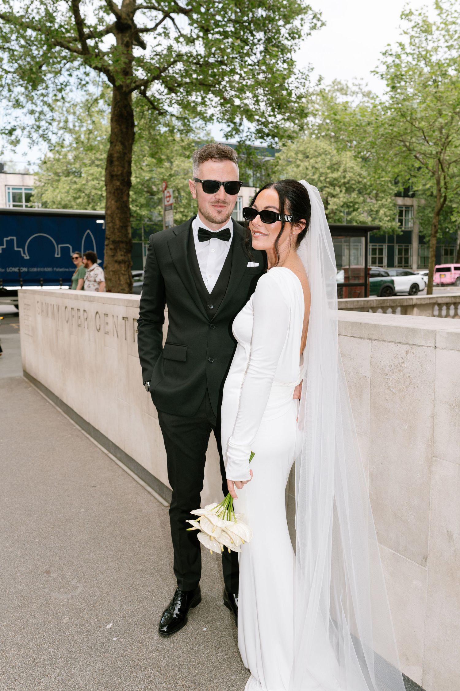 Newlyweds in sunglasses walking along concrete wall on sunny spring day.