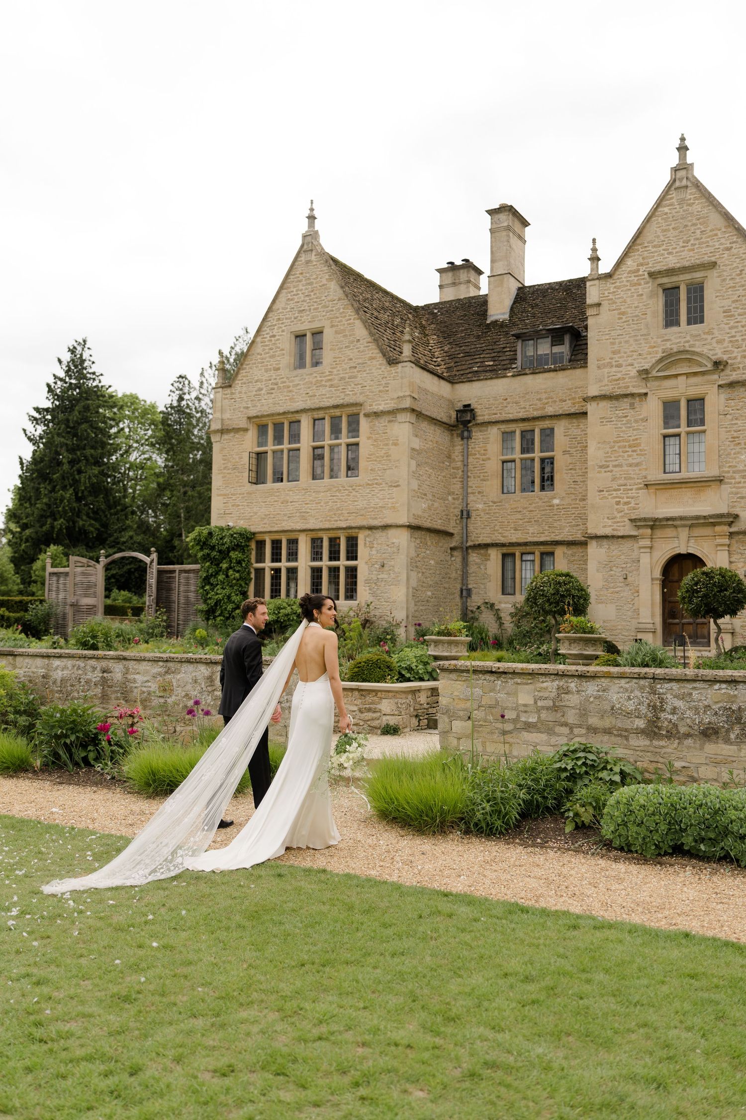 Bride and groom pose in front of a grand Cotswold stone manor house surrounded by manicured gardens.