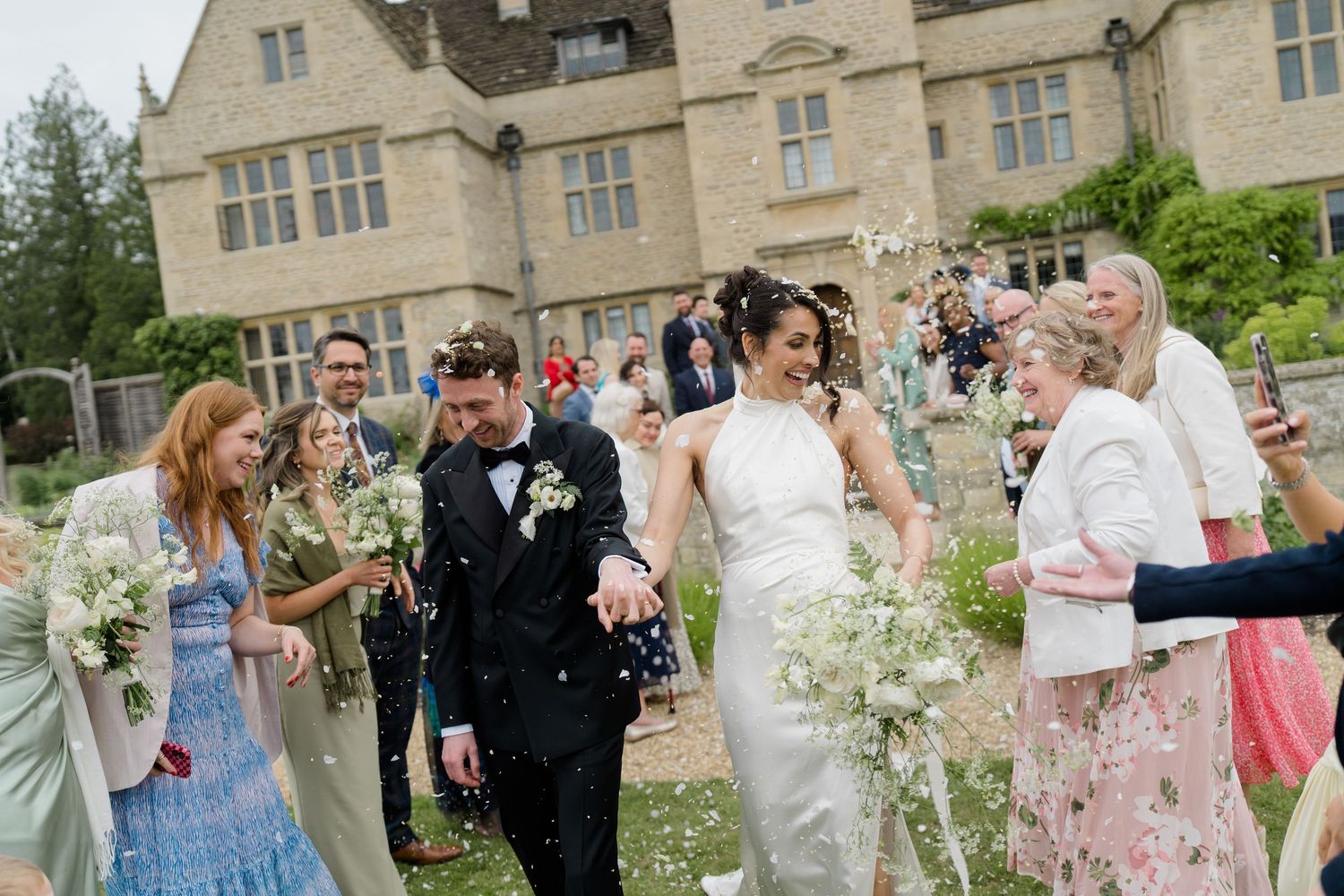 Newlyweds walk through confetti shower outside historic stone manor surrounded by cheering guests.