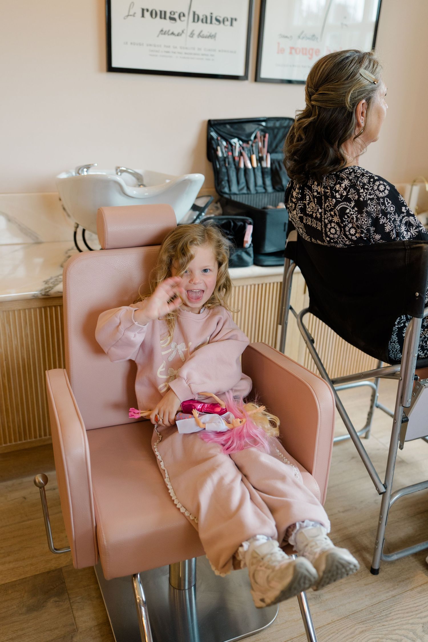 Adorable young girl in pink outfit sitting in salon chair smiling during wedding preparation at beauty salon.