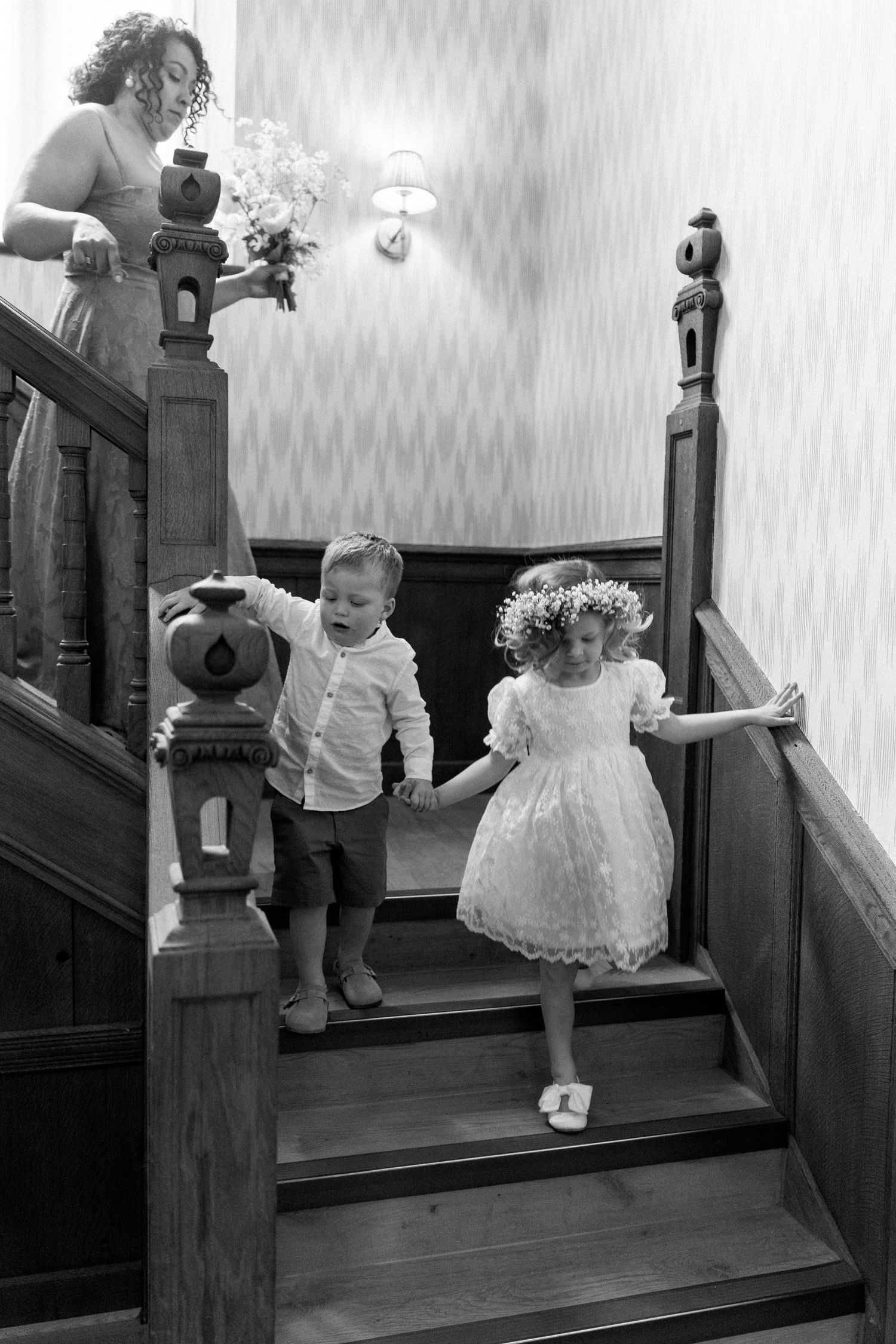 Black and white photo of flower girl and page boy holding hands descending wooden staircase.