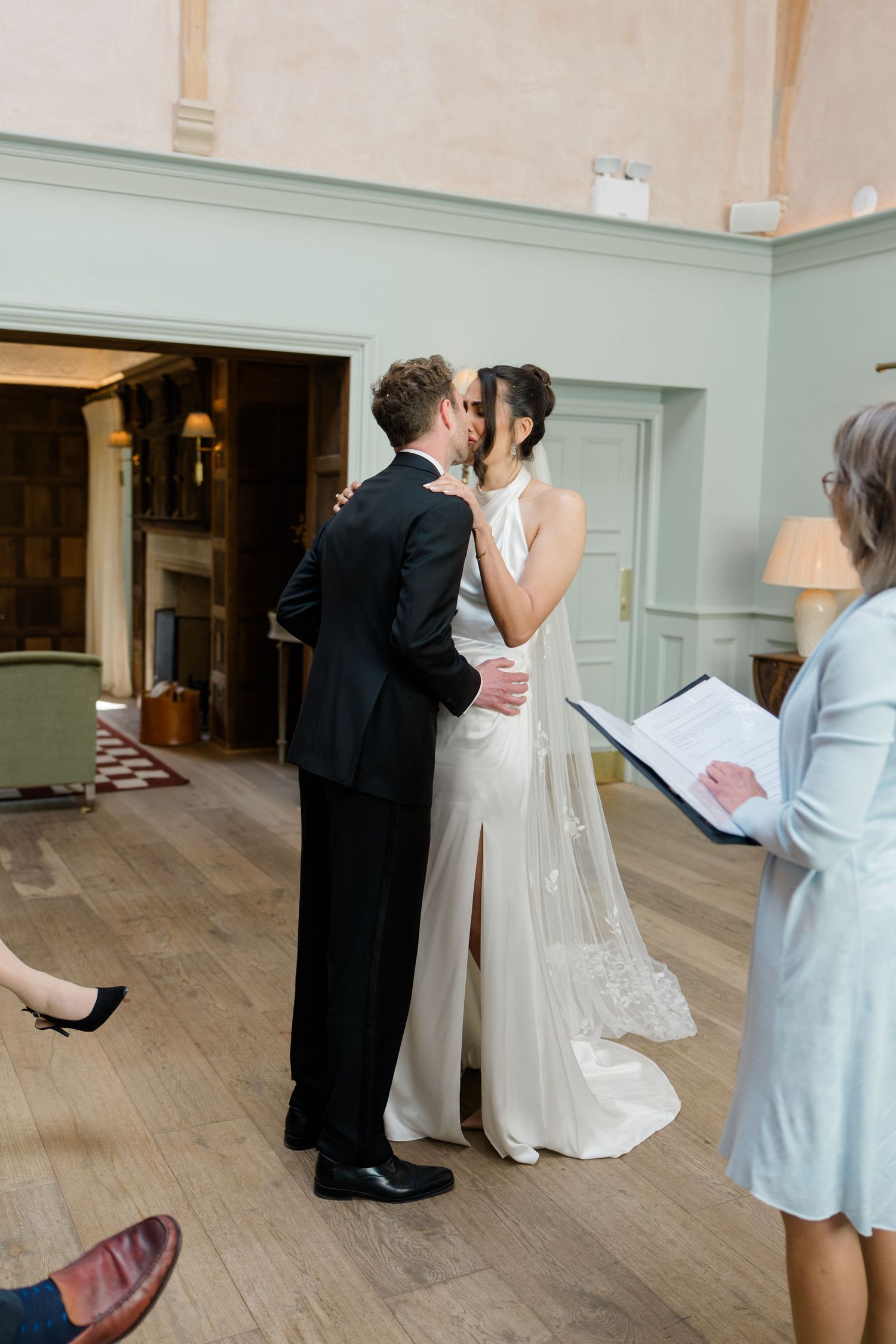 Bride and groom sharing first kiss at elegant indoor wedding ceremony with officiant nearby.