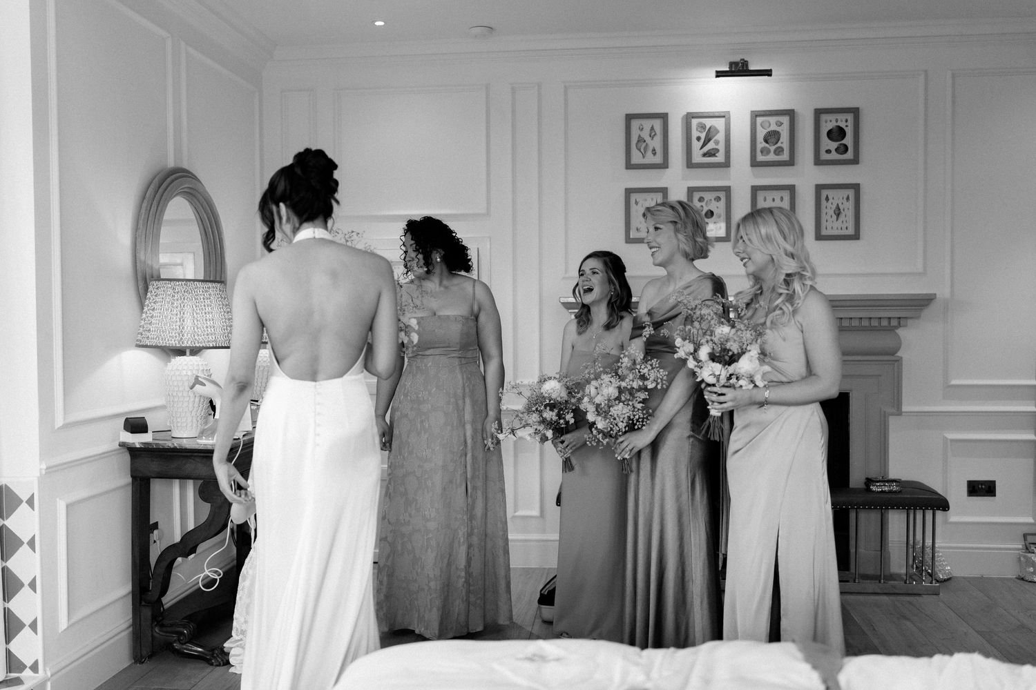 A bride in a backless gown stands with bridesmaids holding bouquets in a elegant room.
