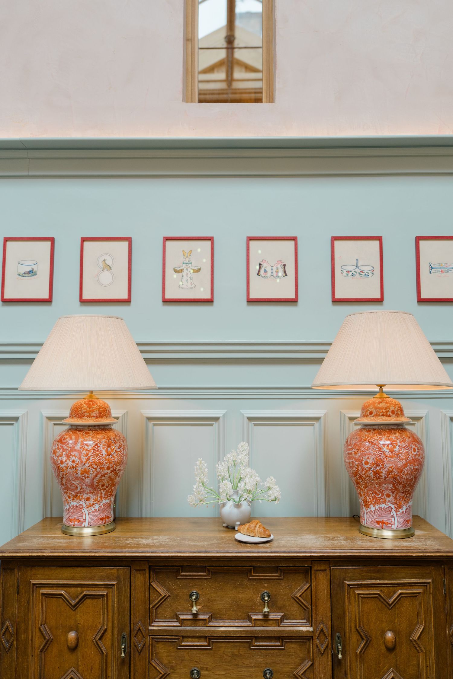 Antique wooden sideboard with two matching orange ceramic table lamps beneath framed botanical prints on blue wall.