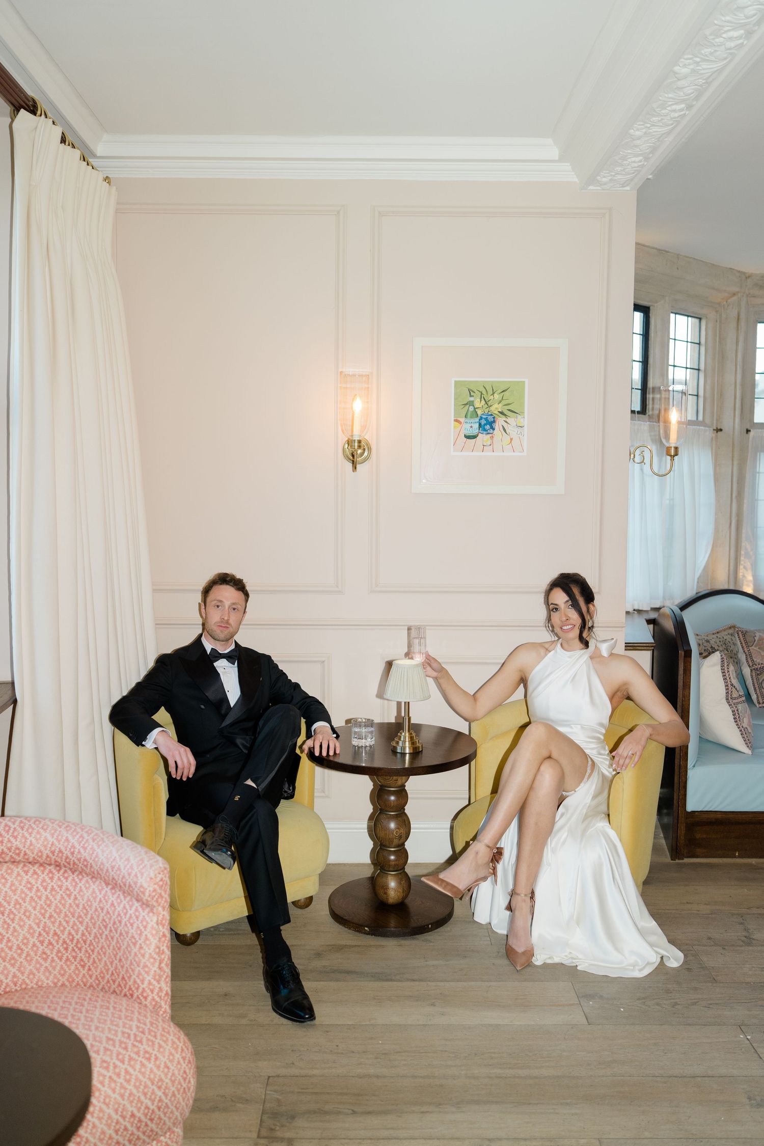 Bride and groom relaxing in elegant yellow armchairs in a softly lit, pastel-toned room.