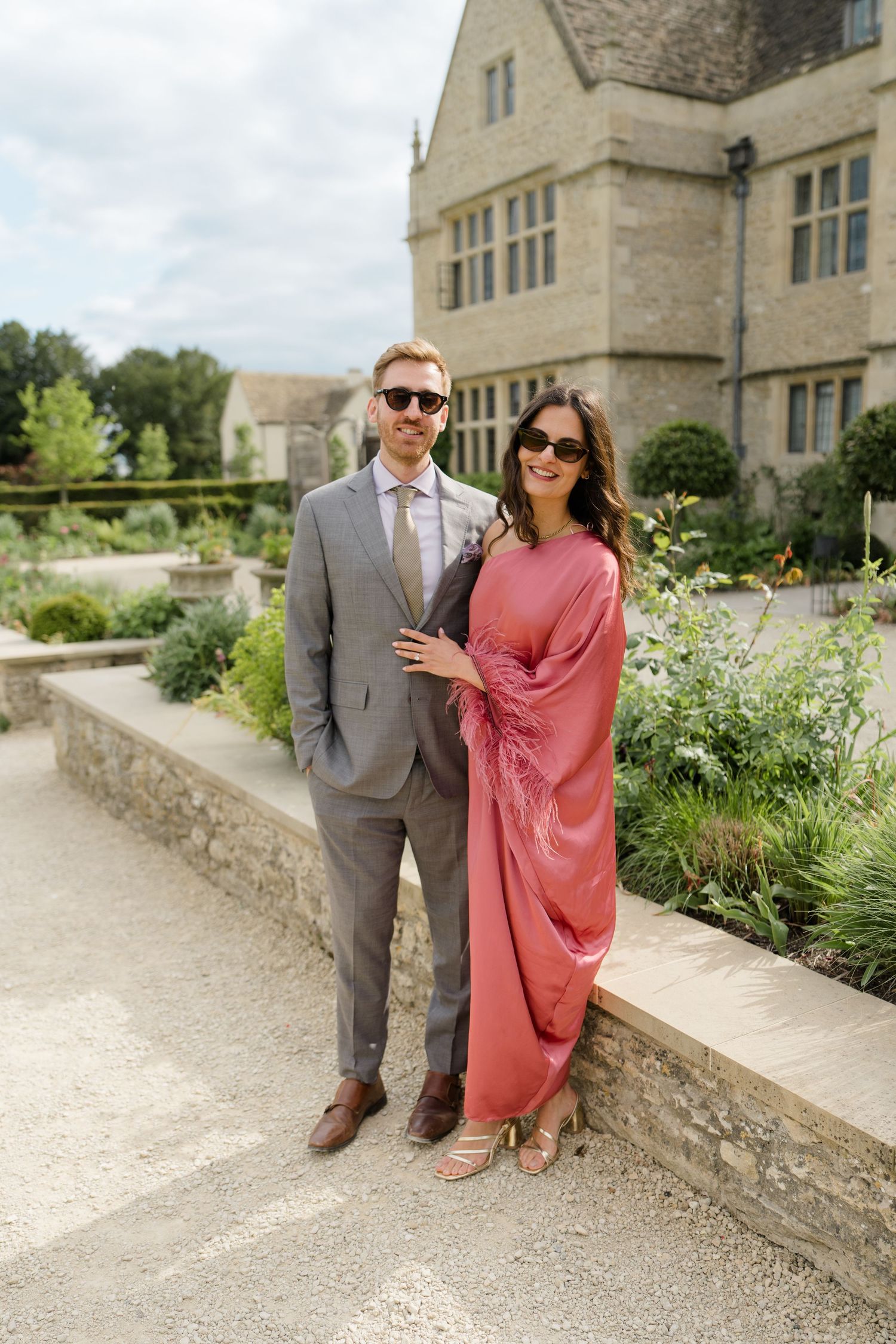 Couple posing in front of a stone manor house, woman in pink feathered dress, man in grey suit with sunglasses.