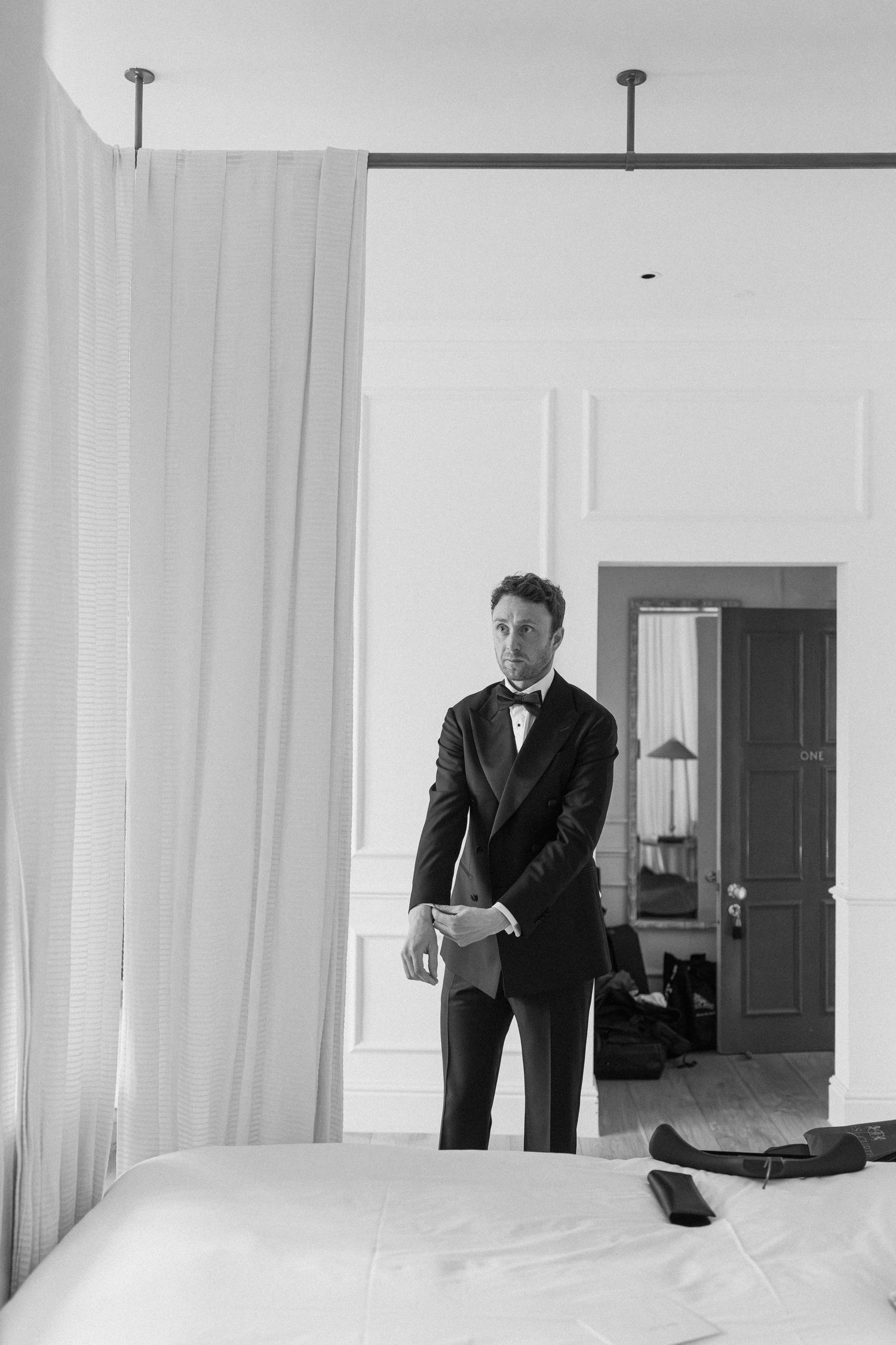 Dapper groom in black tuxedo and bow tie standing in a bright elegant hotel suite.