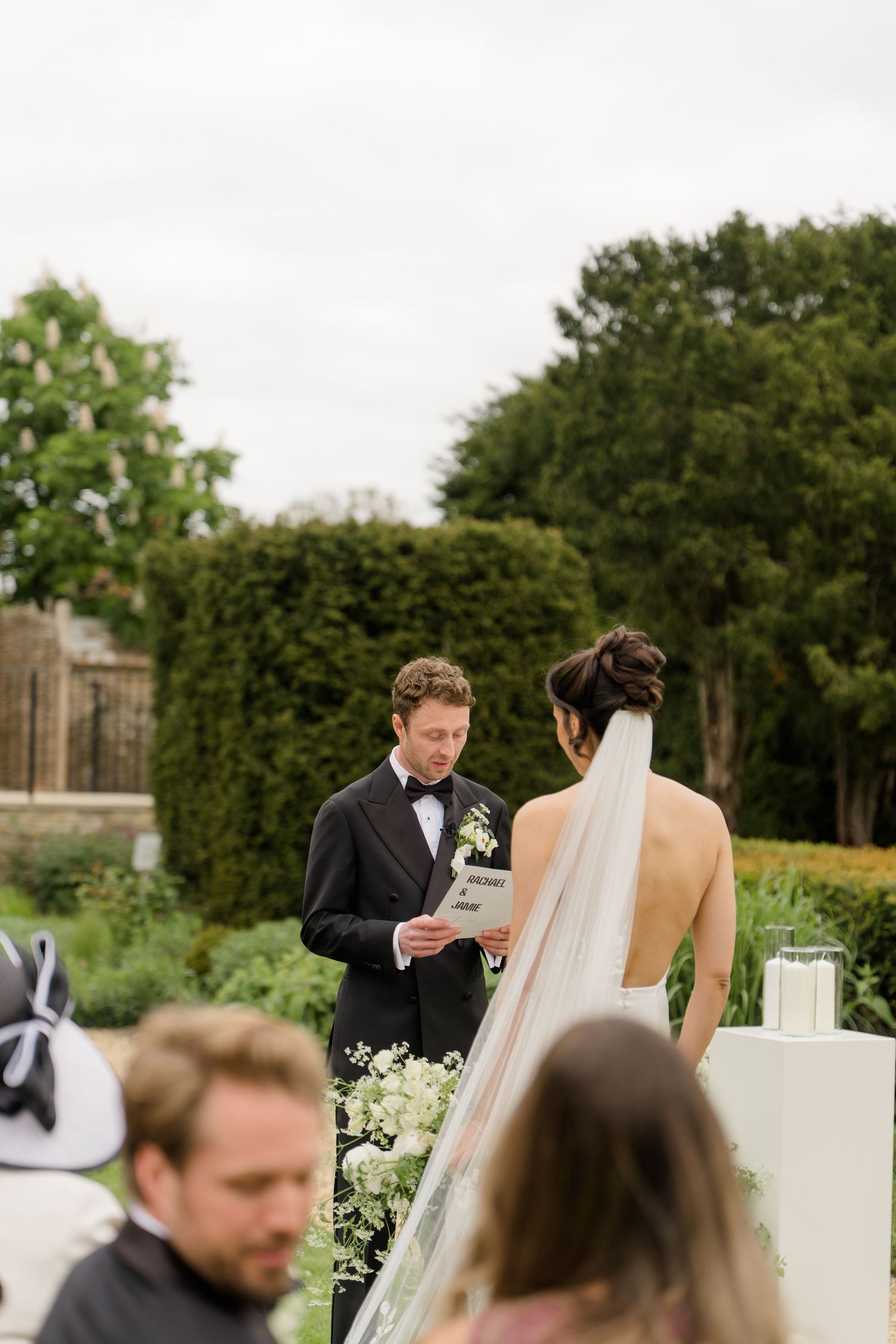 Groom in black tuxedo reads vows to bride in backless wedding dress during outdoor garden ceremony.