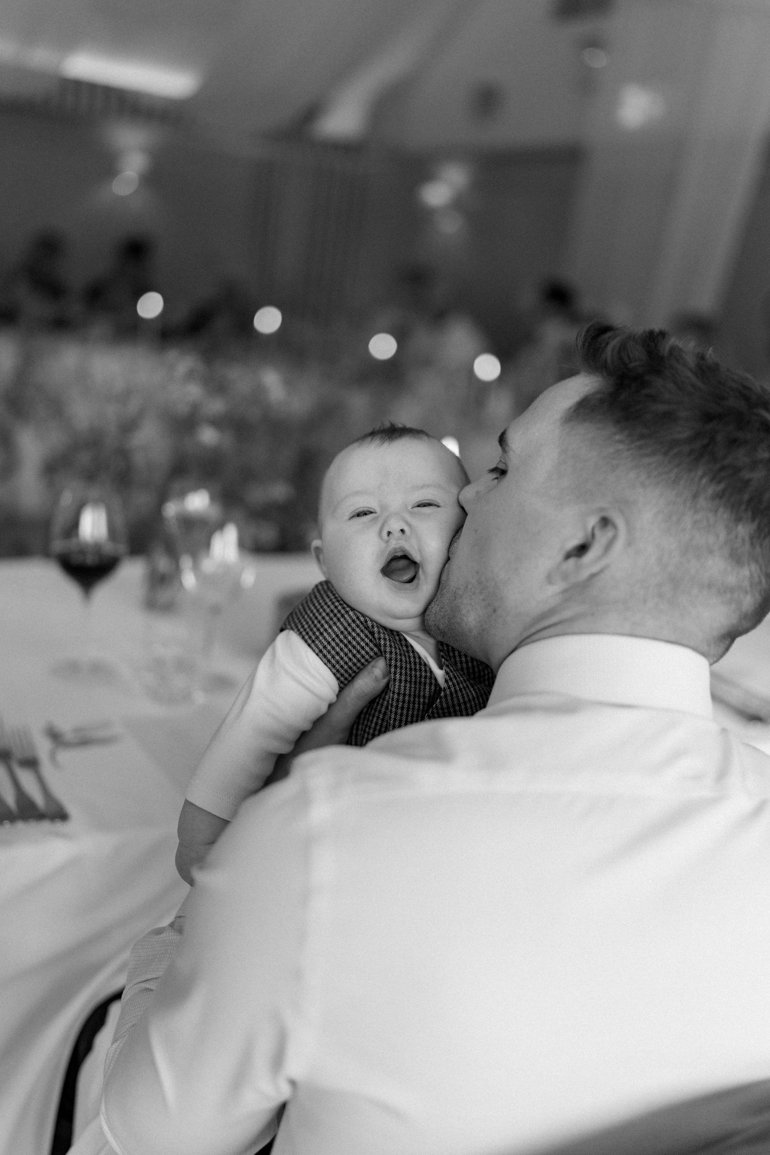 Tender black and white photo of a man kissing a laughing baby at a wedding reception.