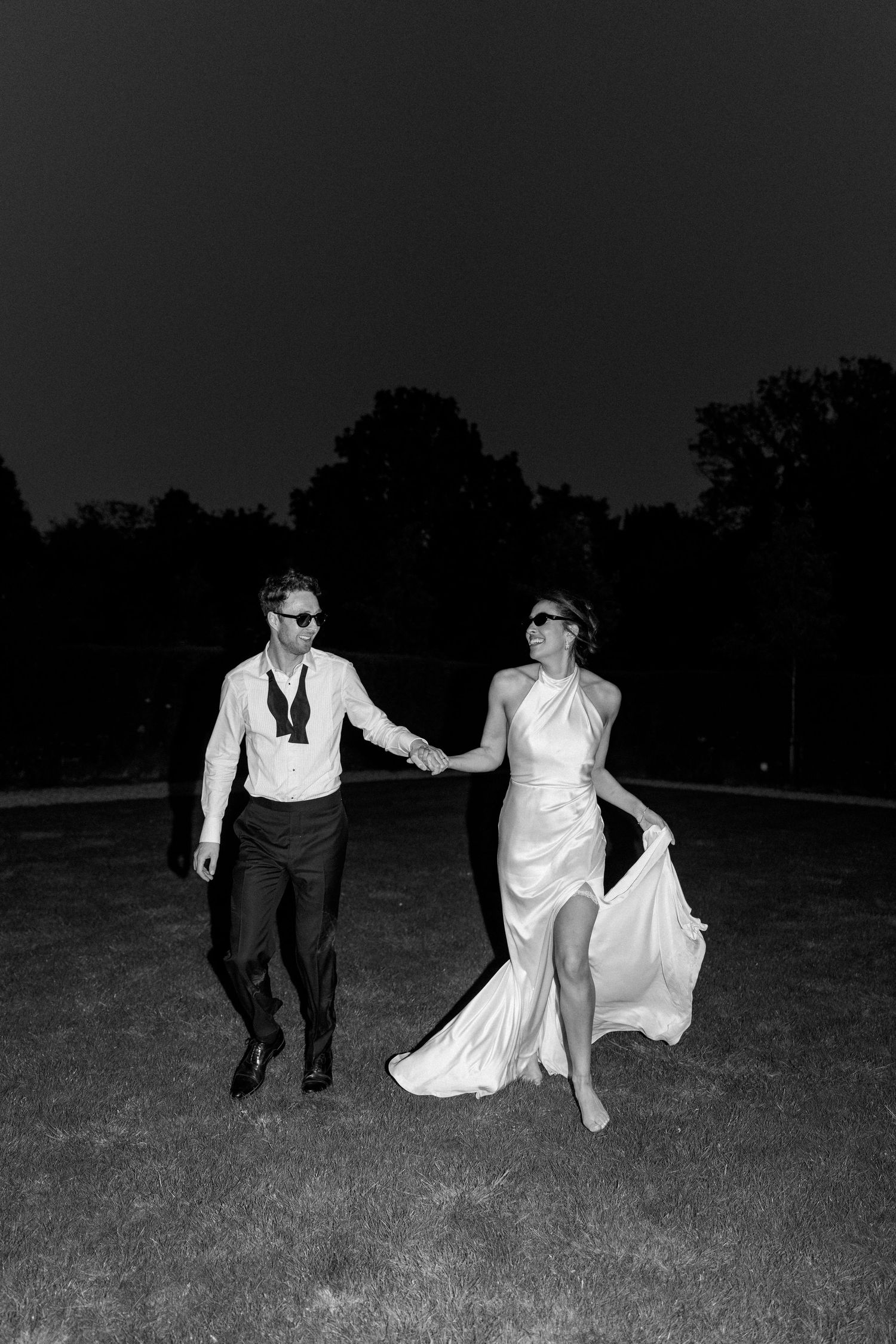 Bride and groom holding hands running across lawn at night in black and white wedding photo.