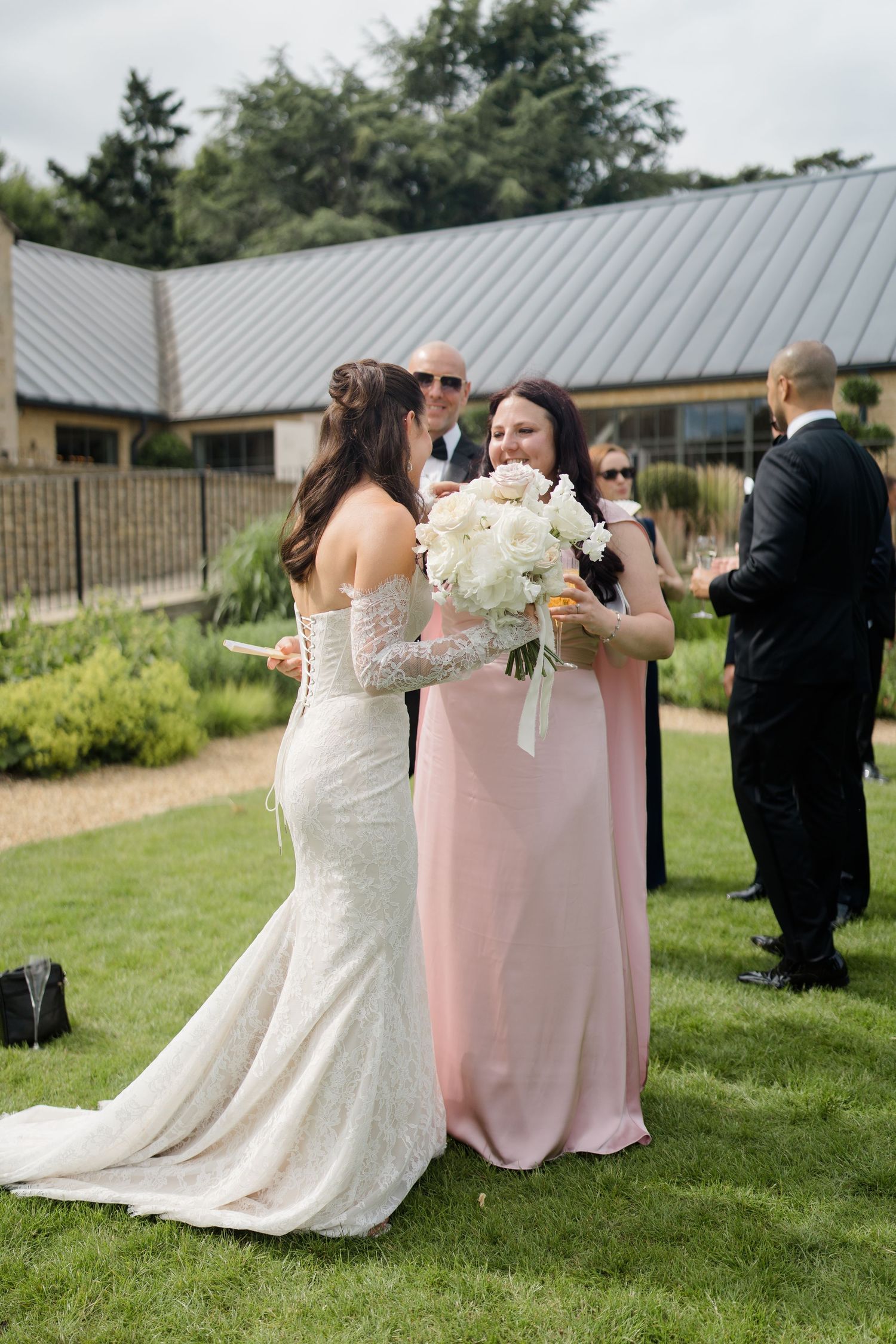 Bride in lace gown holds white peony bouquet chatting with bridesmaid in pink dress outdoors.