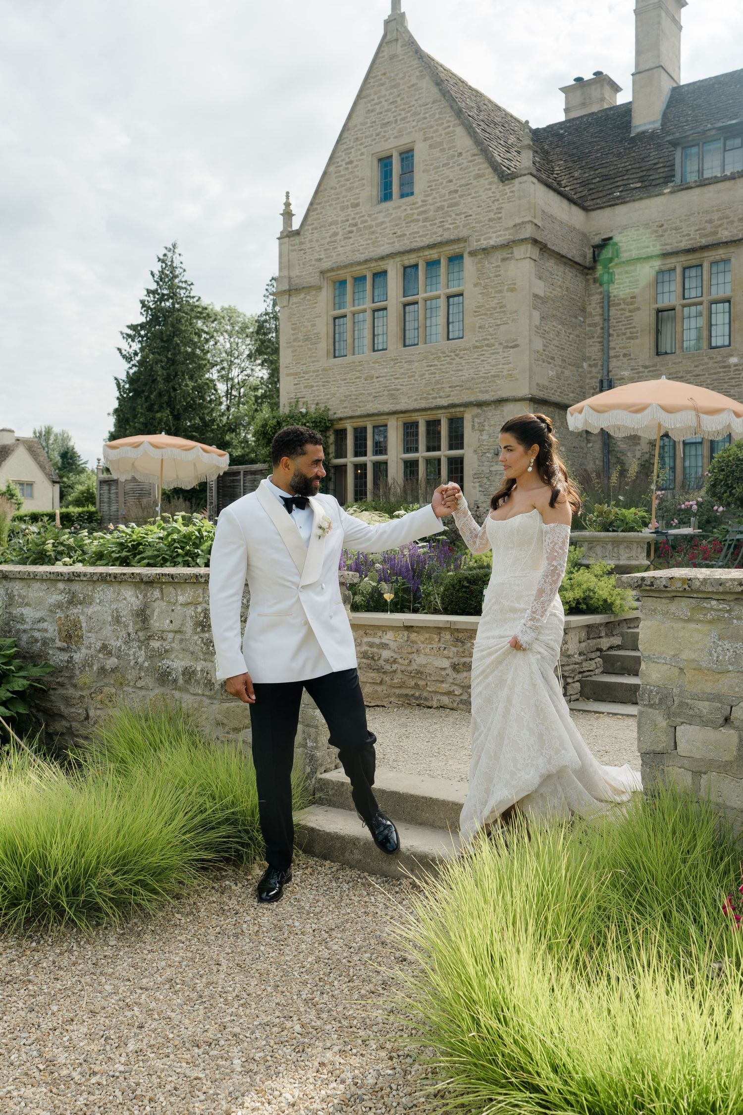 Bride and groom holding hands on garden steps outside elegant stone manor house on their wedding day.
