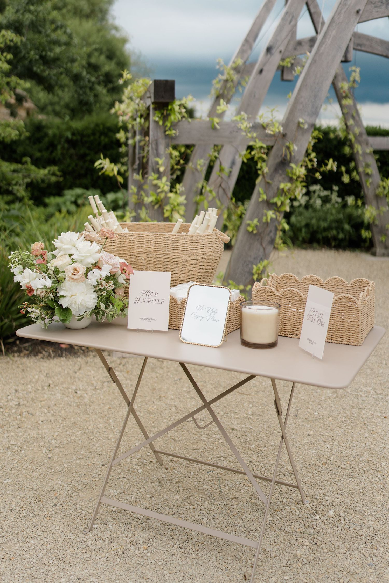 Outdoor wedding welcome table with wicker baskets, floral arrangement, candle and signage.