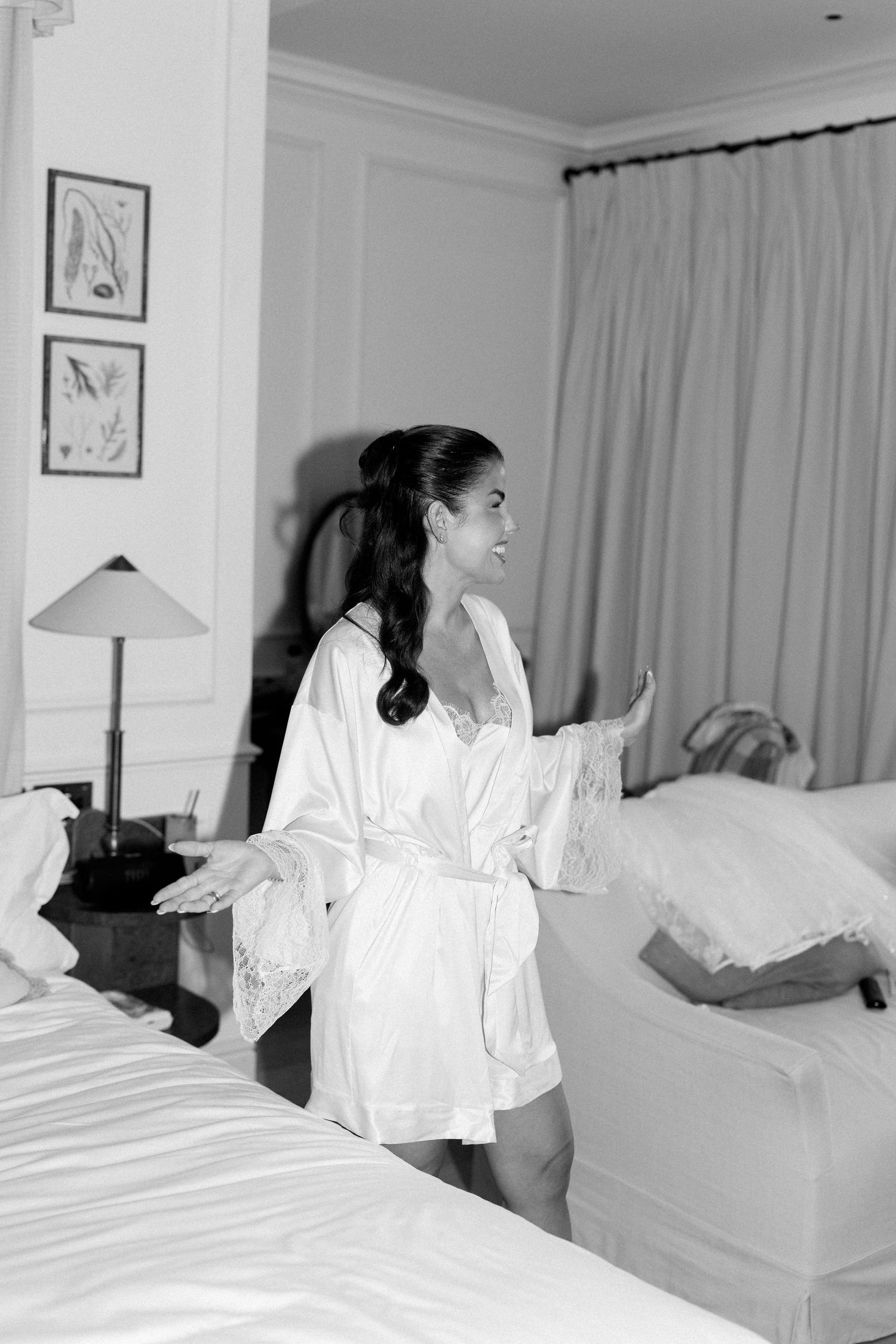 Bride in white silk robe standing between beds in elegant hotel room on wedding morning.