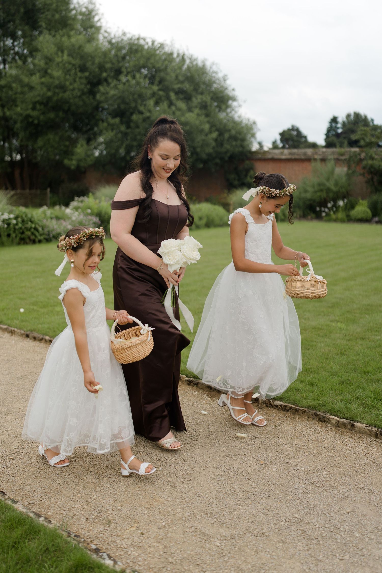 Bridesmaid in dark brown gown walks with two flower girls in white dresses carrying wicker baskets at garden wedding.
