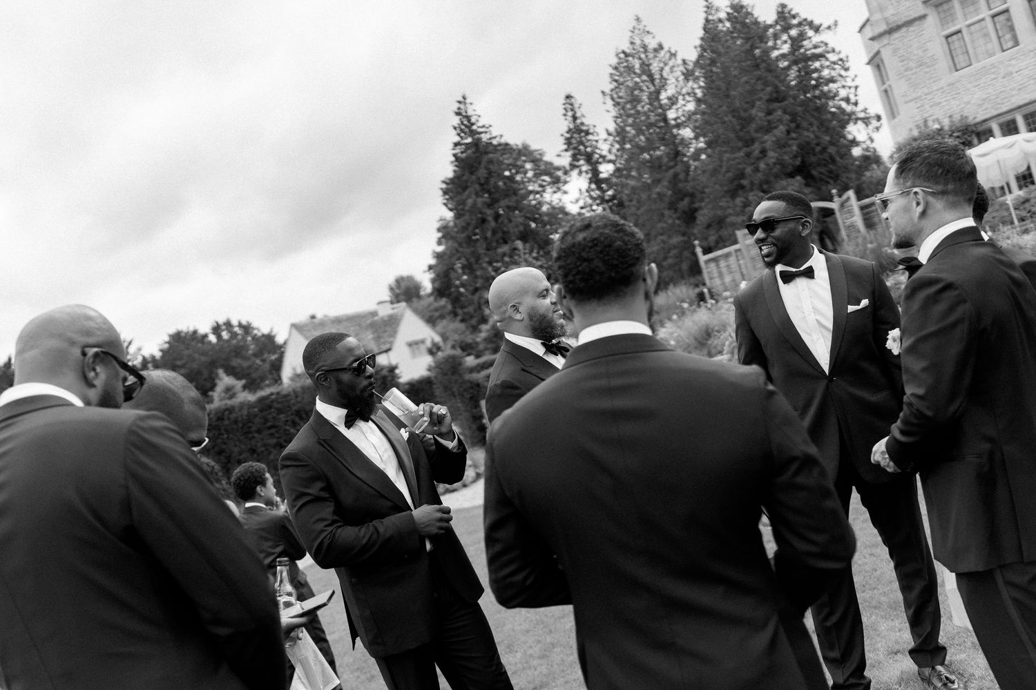 Black and white photo of groomsmen in tuxedos socializing outdoors at a wedding reception.