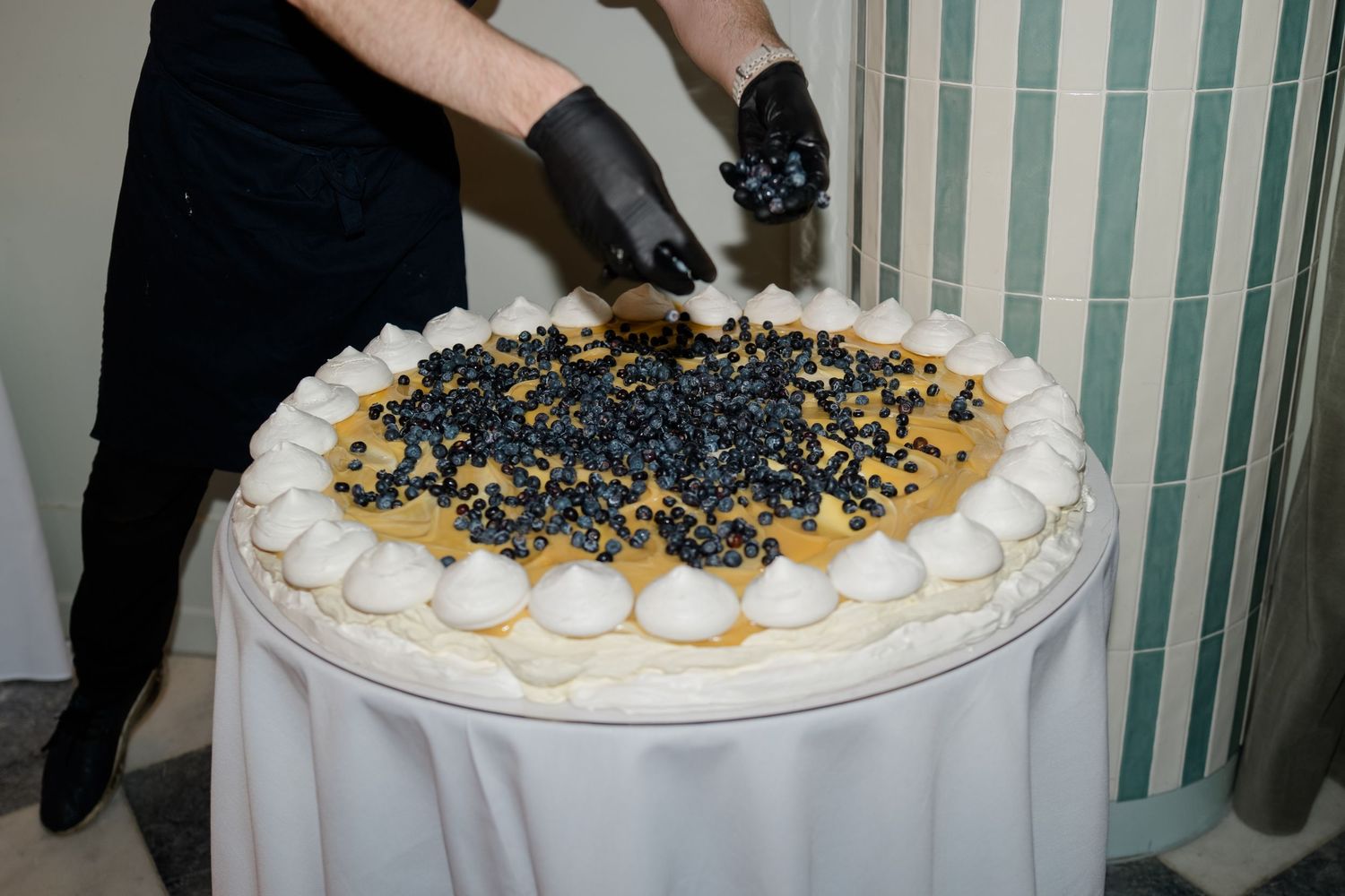 Chef in black gloves decorating large round dessert tart with fresh blueberries at wedding.