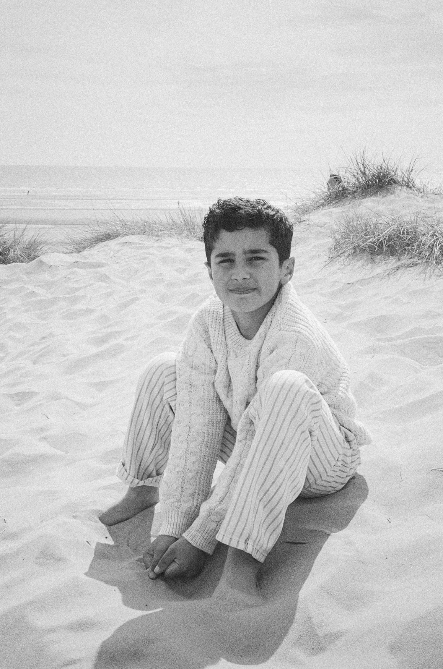 A young boy sits barefoot on sandy beach dunes wearing a cream knit sweater and striped pants in a black and white photo.