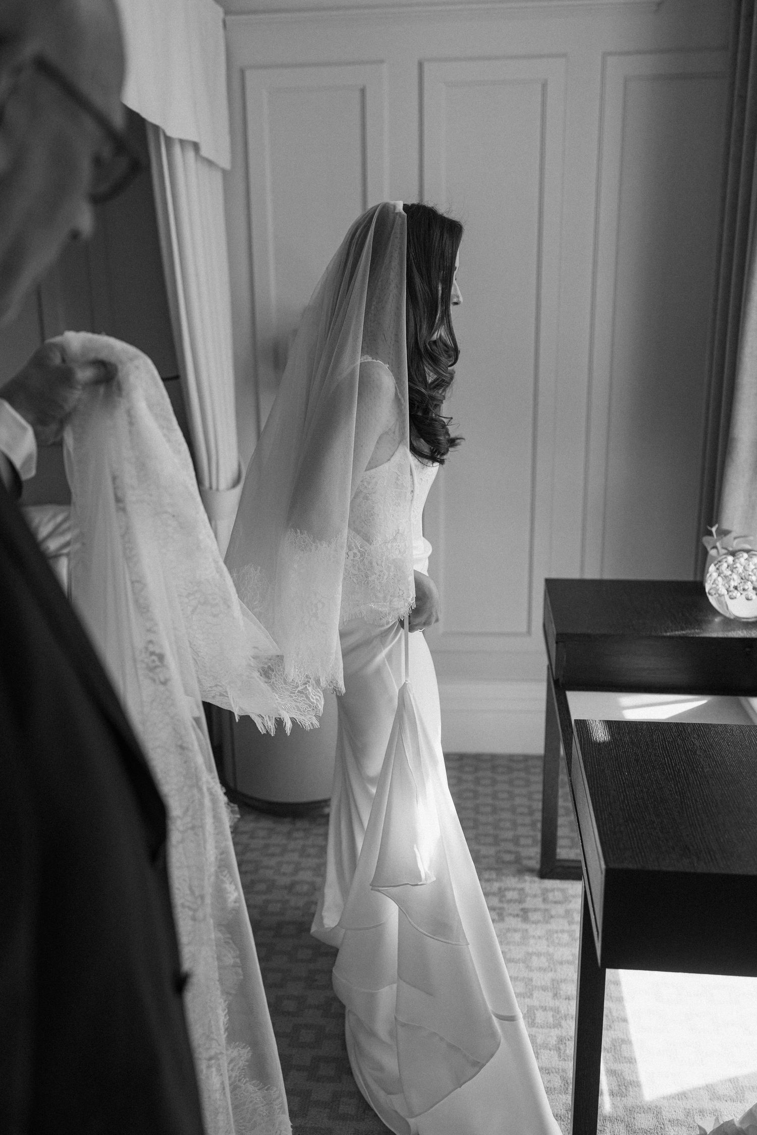 Black and white photo of a bride in a wedding gown and veil getting ready in an elegant room.