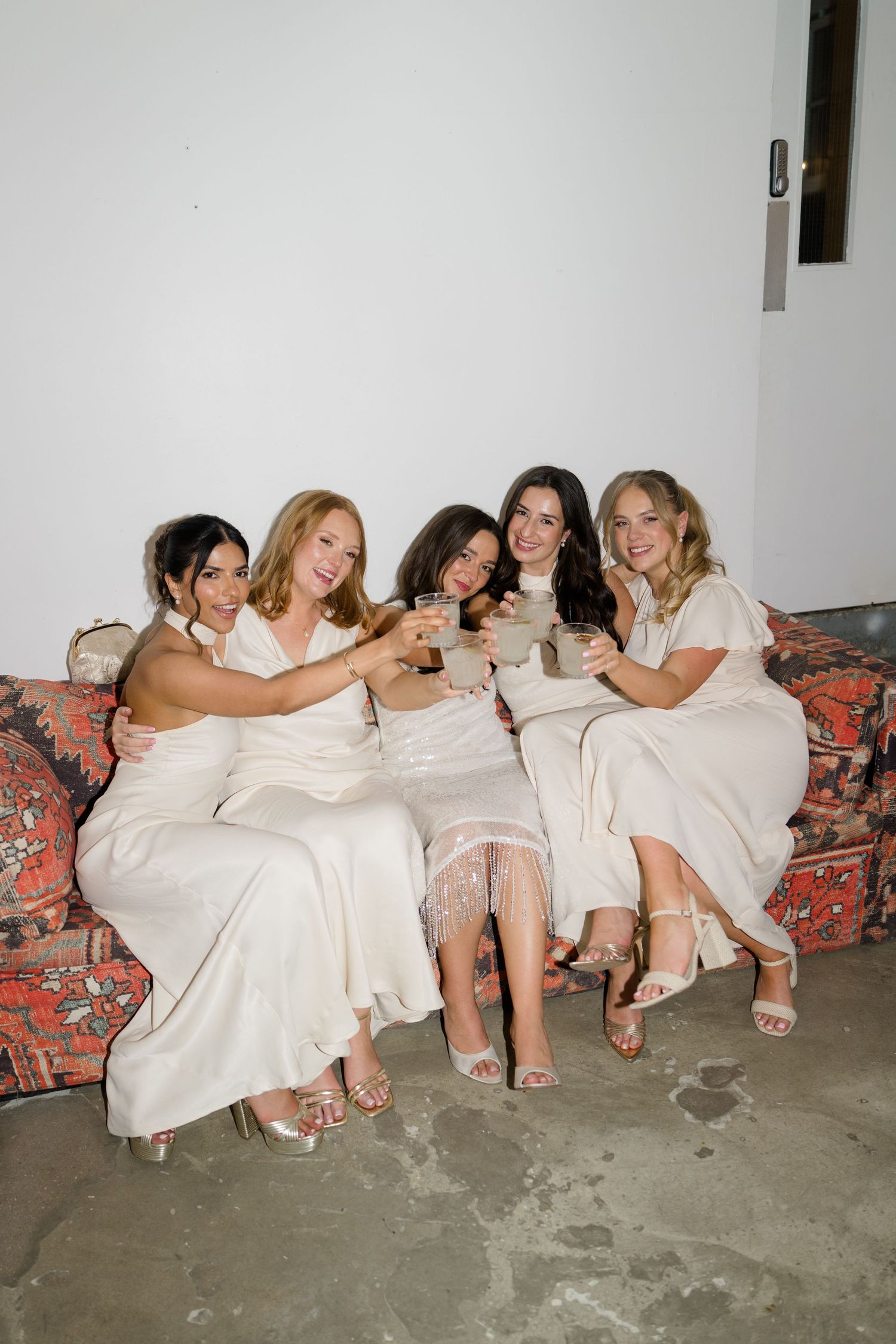 Five women in white dresses toasting with drinks while seated on a floral couch.