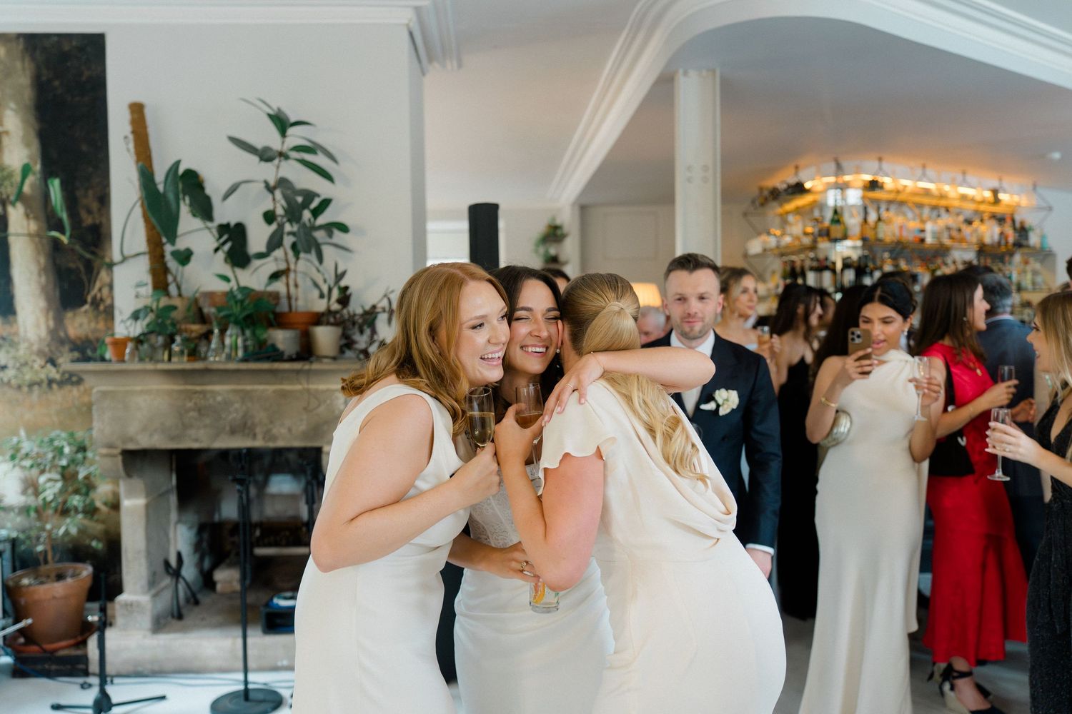 Bride receives warm hugs from two guests in white dresses at a bright wedding reception venue.