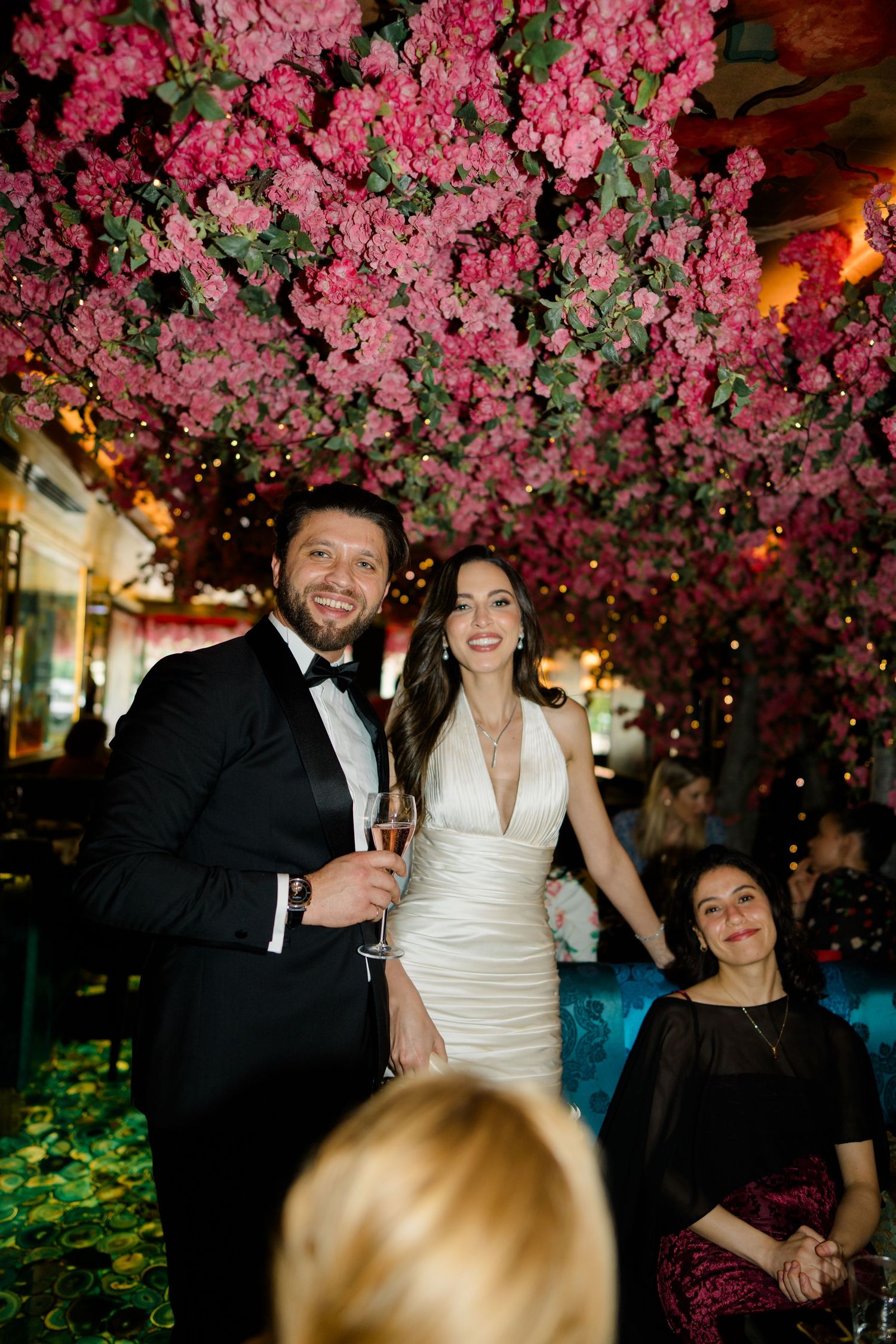 Couple in formal attire smiling under pink floral ceiling decor at elegant wedding reception venue.
