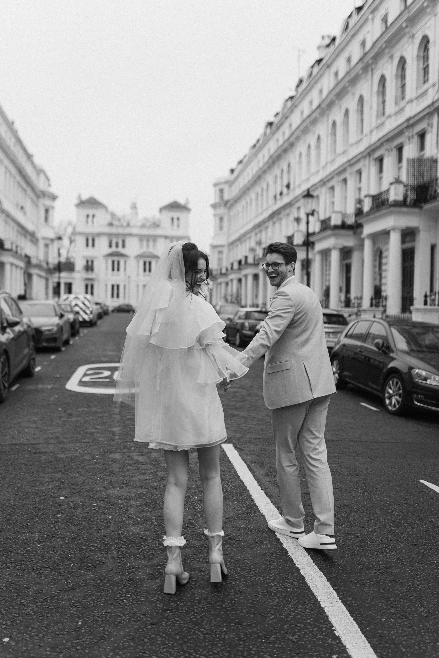 Black and white photo of a bride and groom holding hands on a London street, both looking back at the camera.