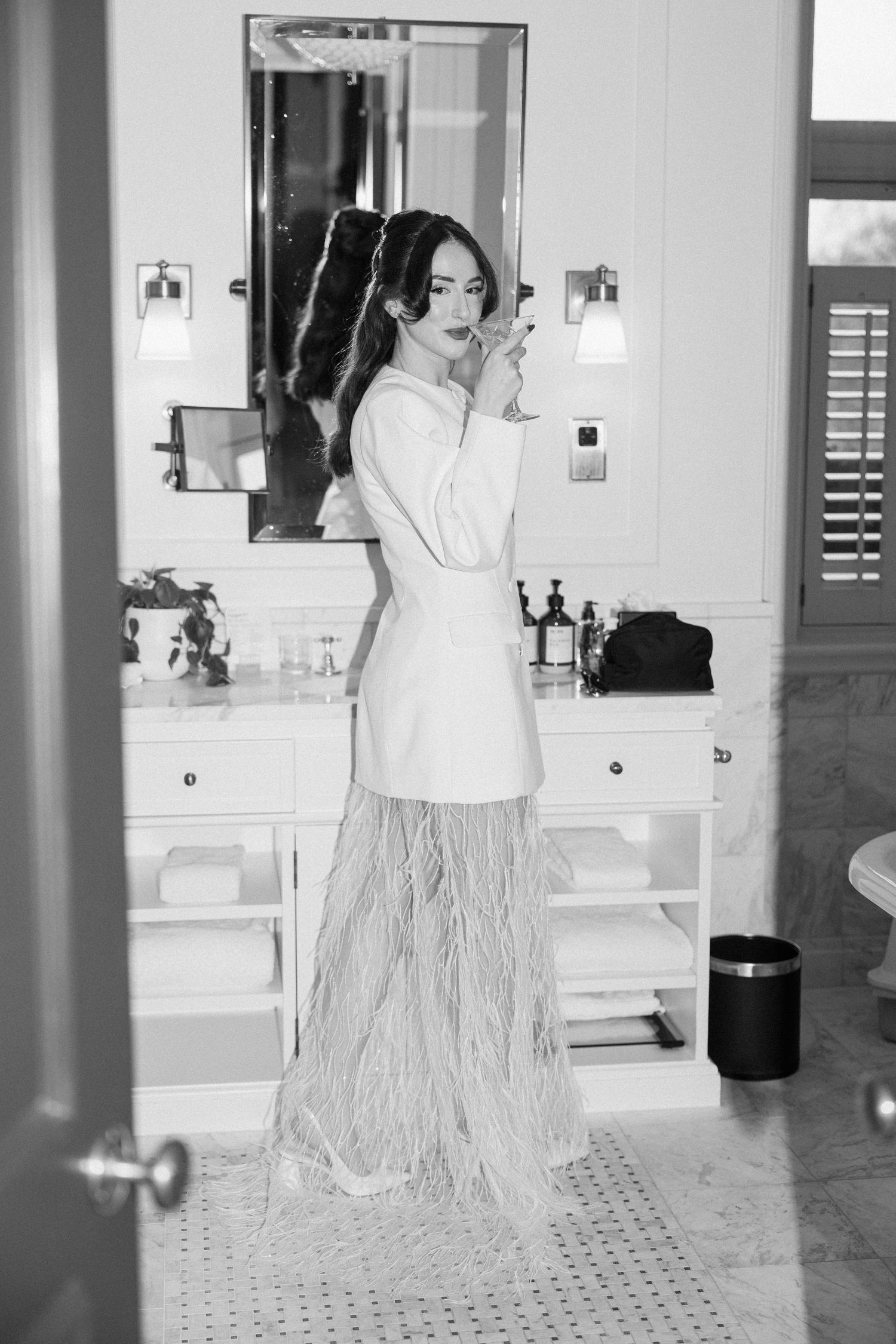 Bride in white blazer and feathered skirt applying lipstick in an elegant bathroom mirror.