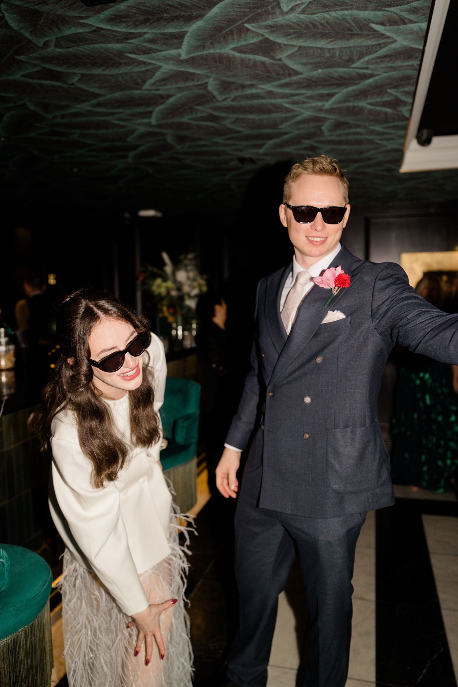 Bride in white blazer and feather skirt poses with groom in navy suit wearing sunglasses in stylish dark interior venue.