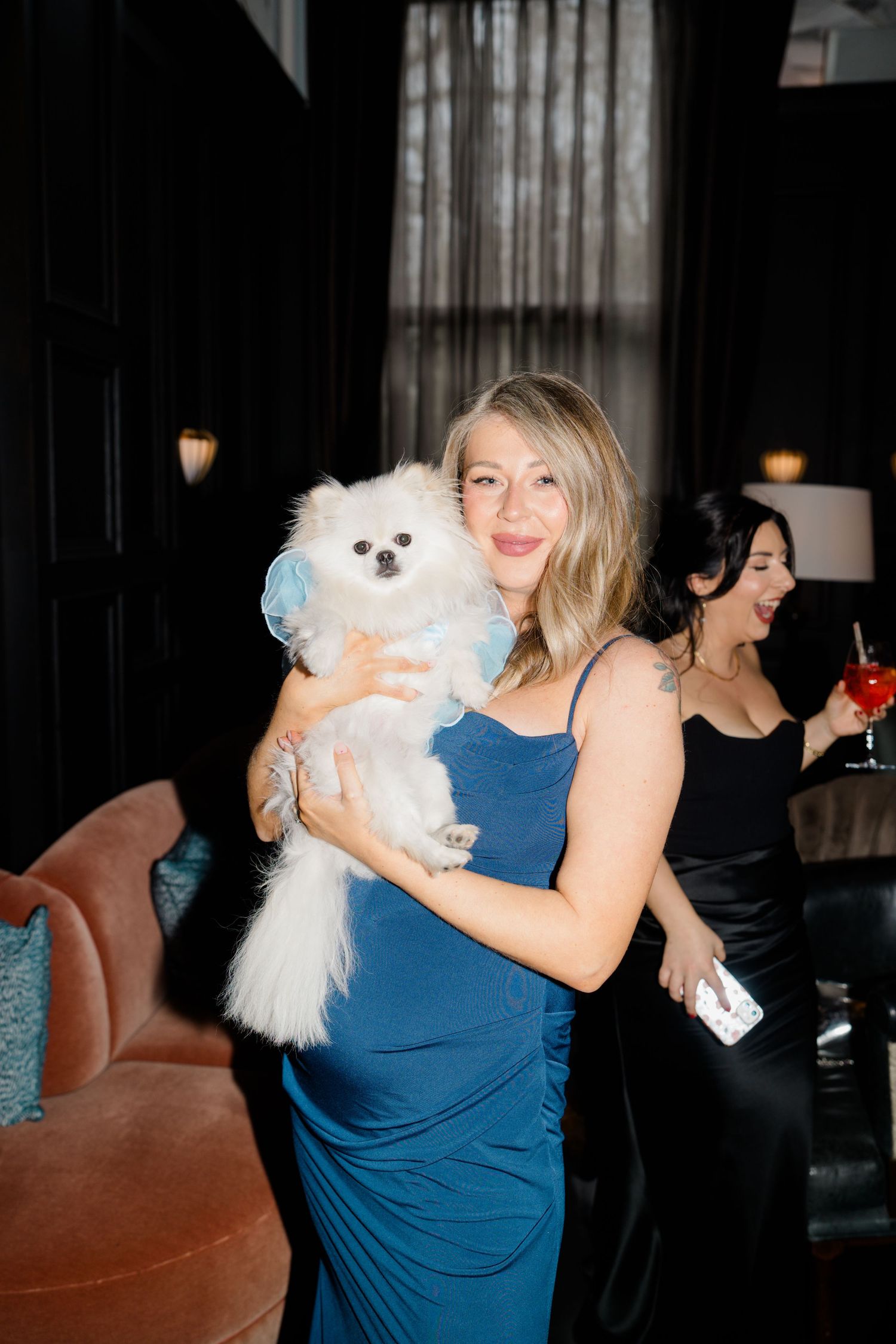 Woman in blue dress holding a white Pomeranian dog at an elegant evening party.