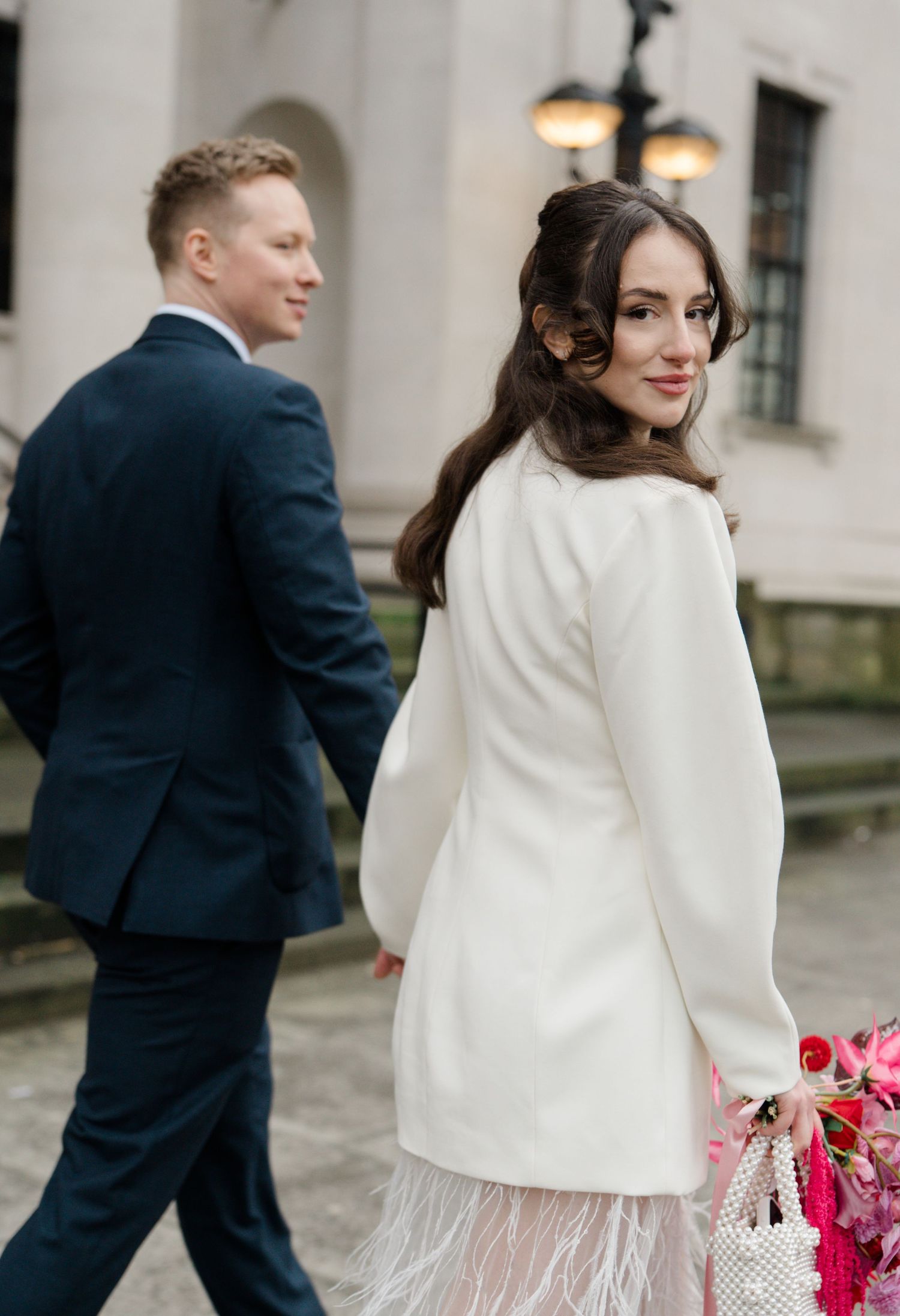 Bride in white feather hem dress glances back while groom in navy suit walks beside her outdoors.