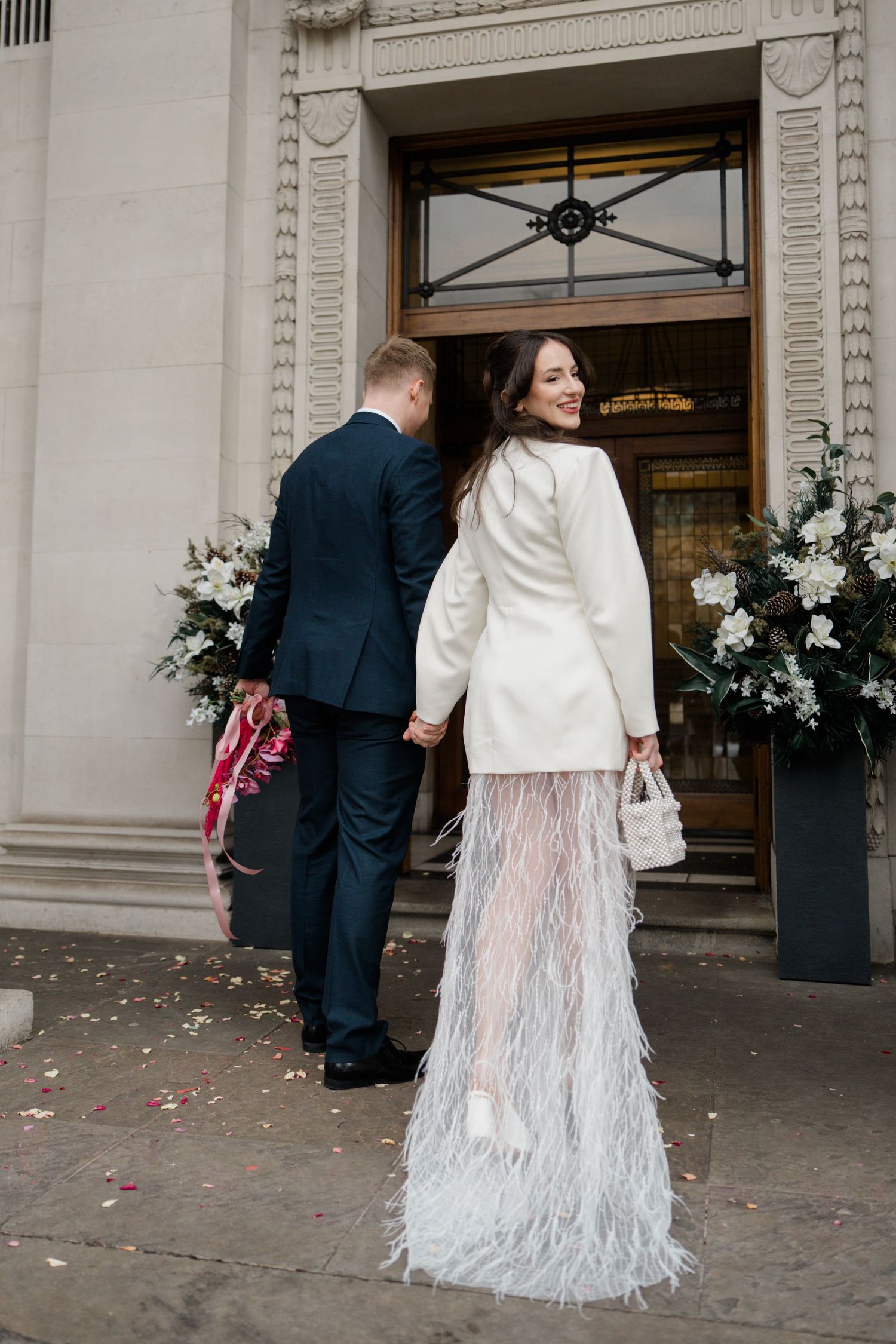 Bride in white feathered skirt and groom in navy suit entering wedding venue doors.