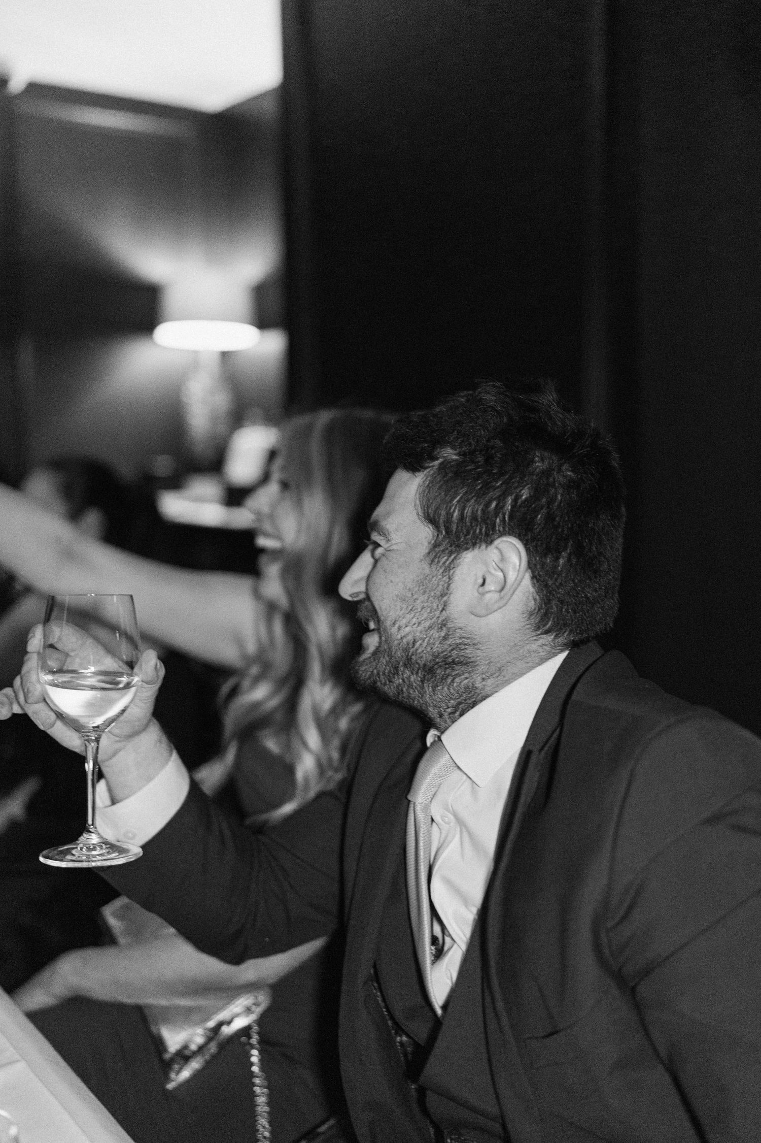 Black and white photo of a bearded man laughing and raising a wine glass at a wedding.