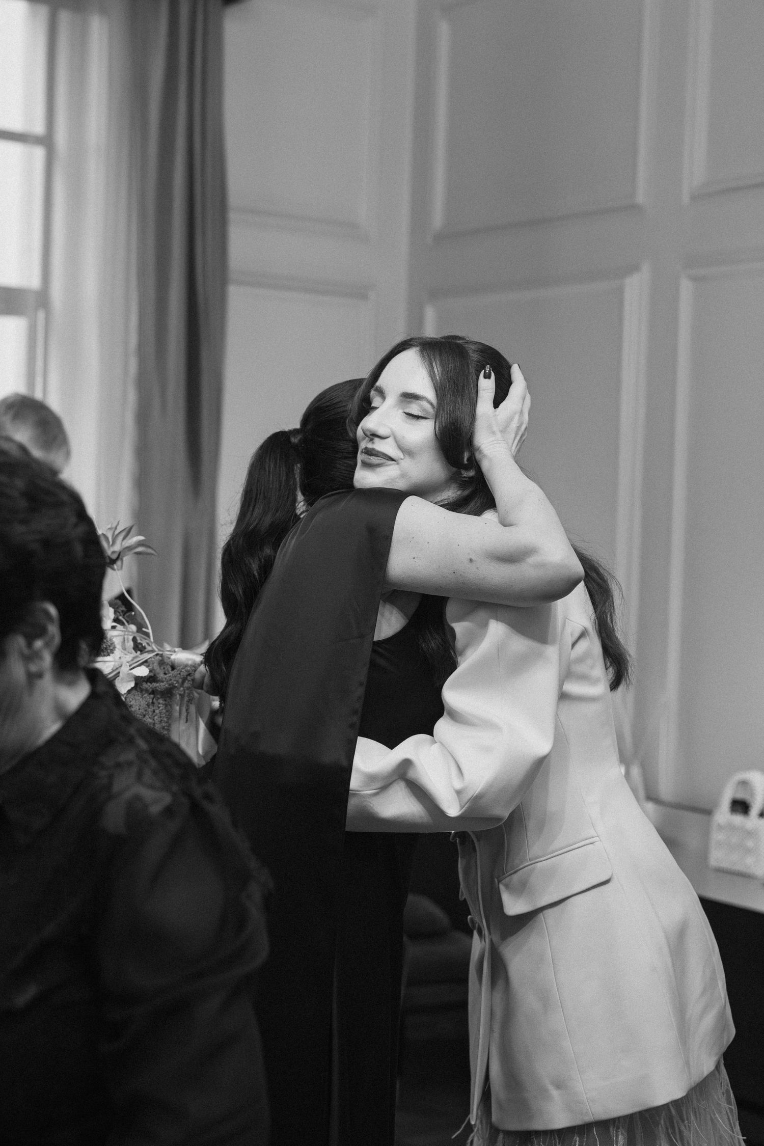 Black and white photo of a bride in a white blazer sharing a joyful hug with a wedding guest.