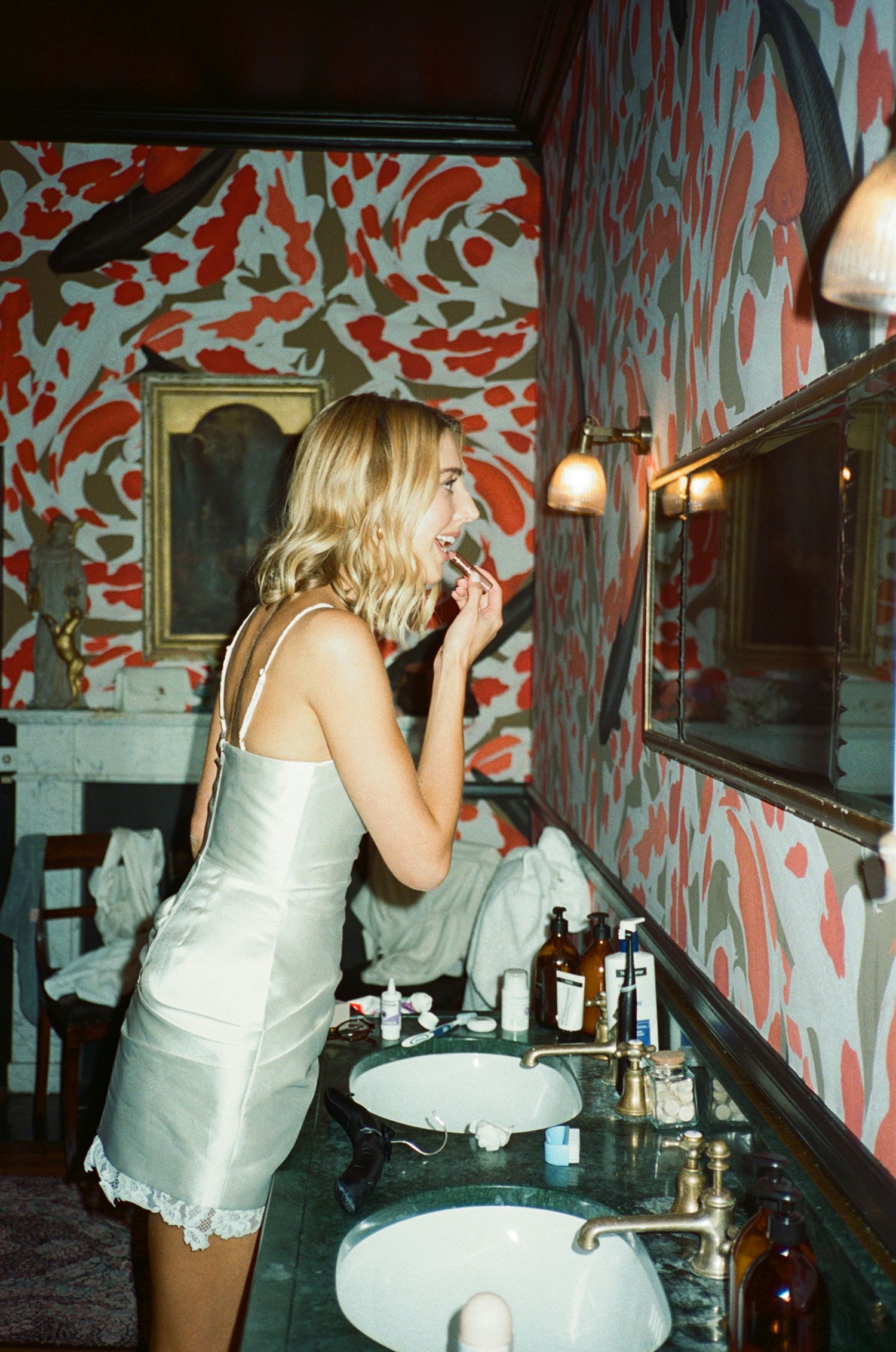 A blonde woman in a silver slip dress applies lipstick at a vintage bathroom vanity with bold red floral wallpaper.