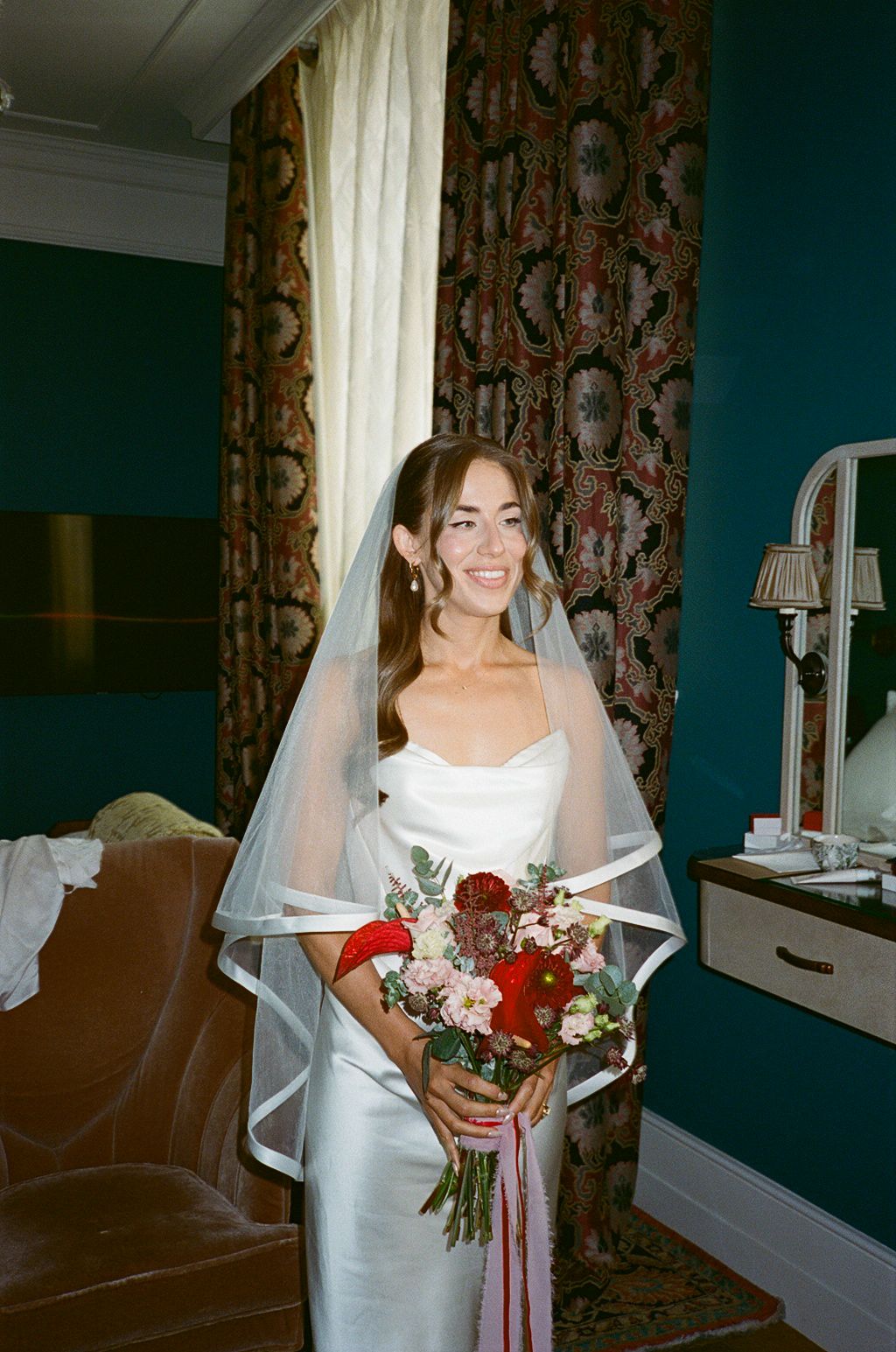 Smiling bride in white strapless gown and veil holding red and pink bouquet in elegant teal room.