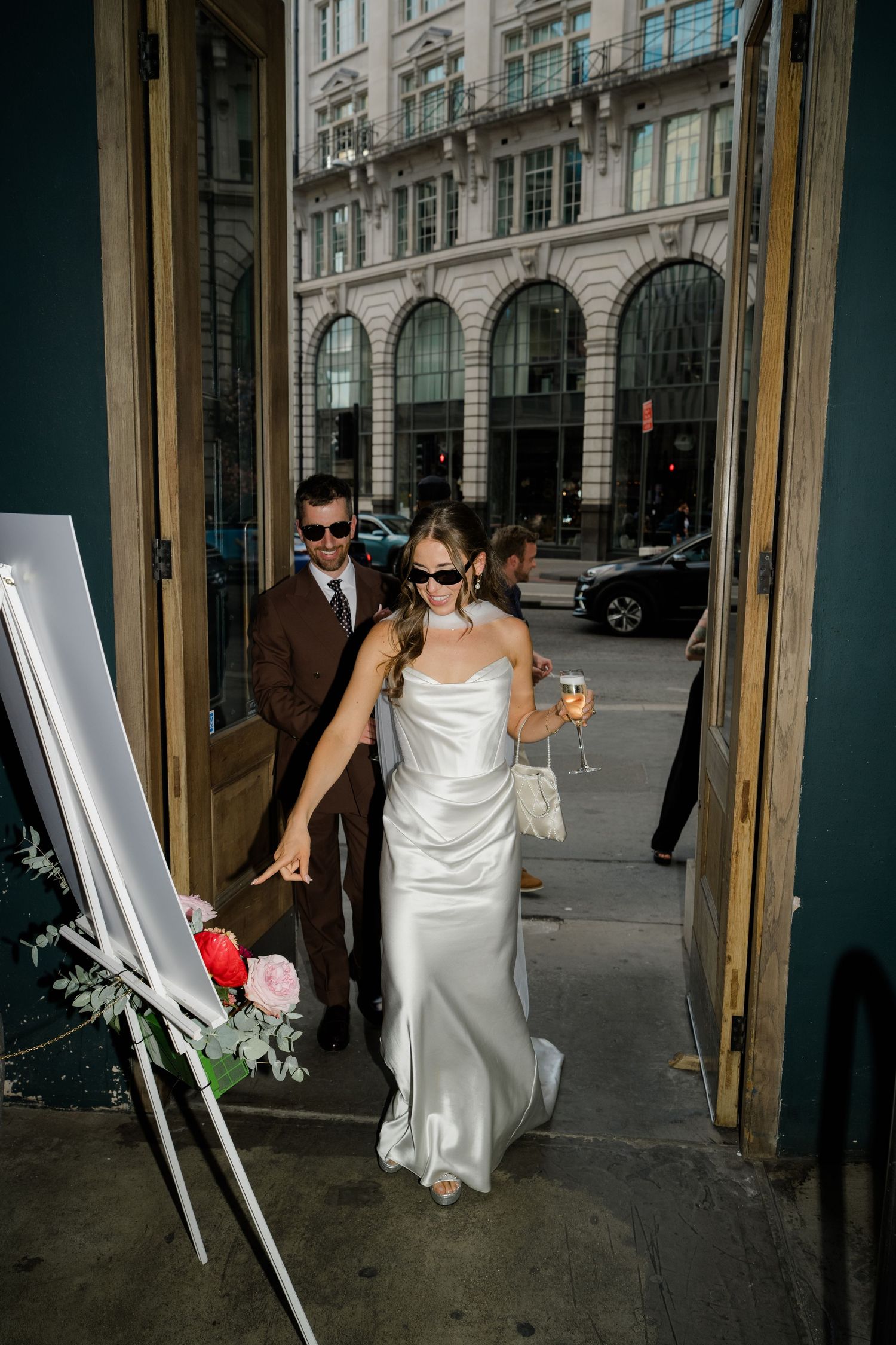 Bride in white satin dress with champagne and groom in brown suit enter venue doorway.