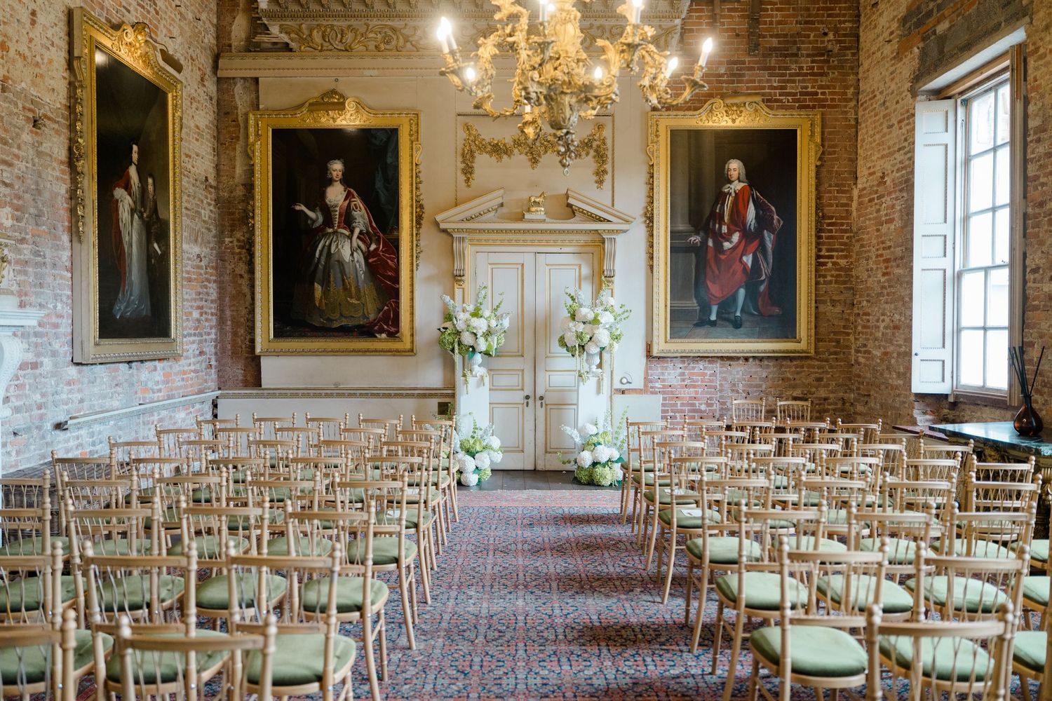 Elegant wedding ceremony setup in historic venue with gold chairs, floral arch, chandelier, and ornate portrait paintings.