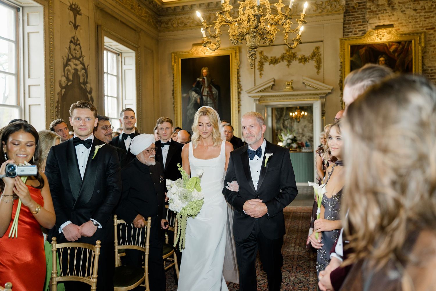 Bride walks down the aisle with escort in elegant gold ballroom wedding ceremony with guests in formal attire.