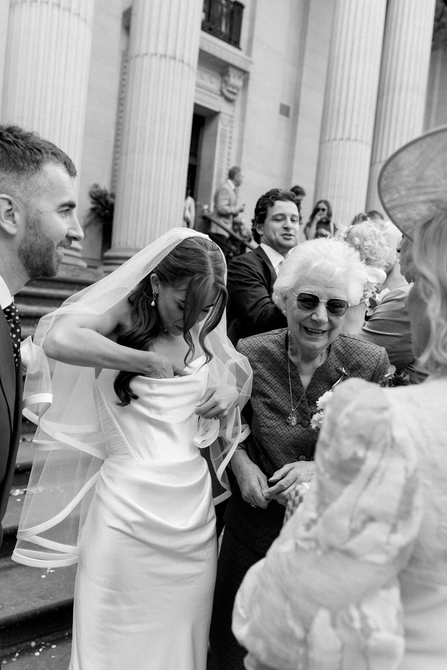 Bride greeting elderly guest outside wedding venue columns in black and white.