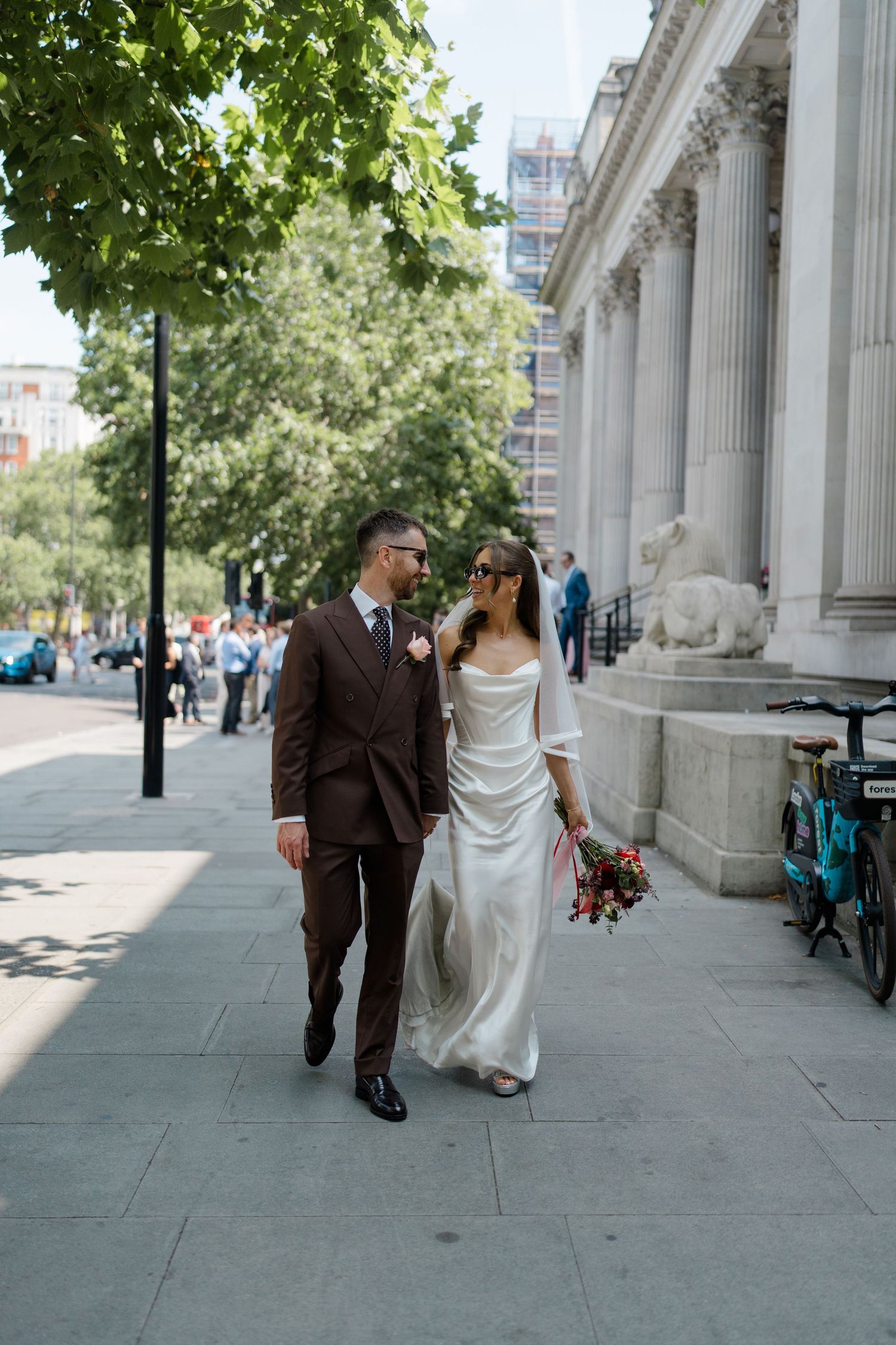Bride and groom walking hand in hand on sunny city sidewalk beside neoclassical building columns.