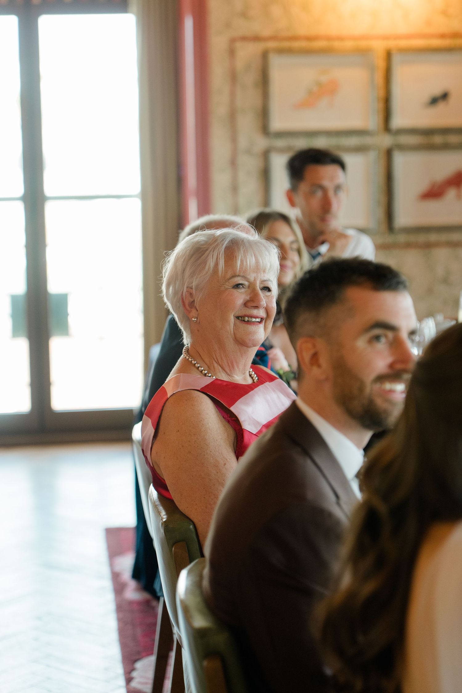 Smiling woman in red dress seated among wedding guests during indoor reception ceremony speech.