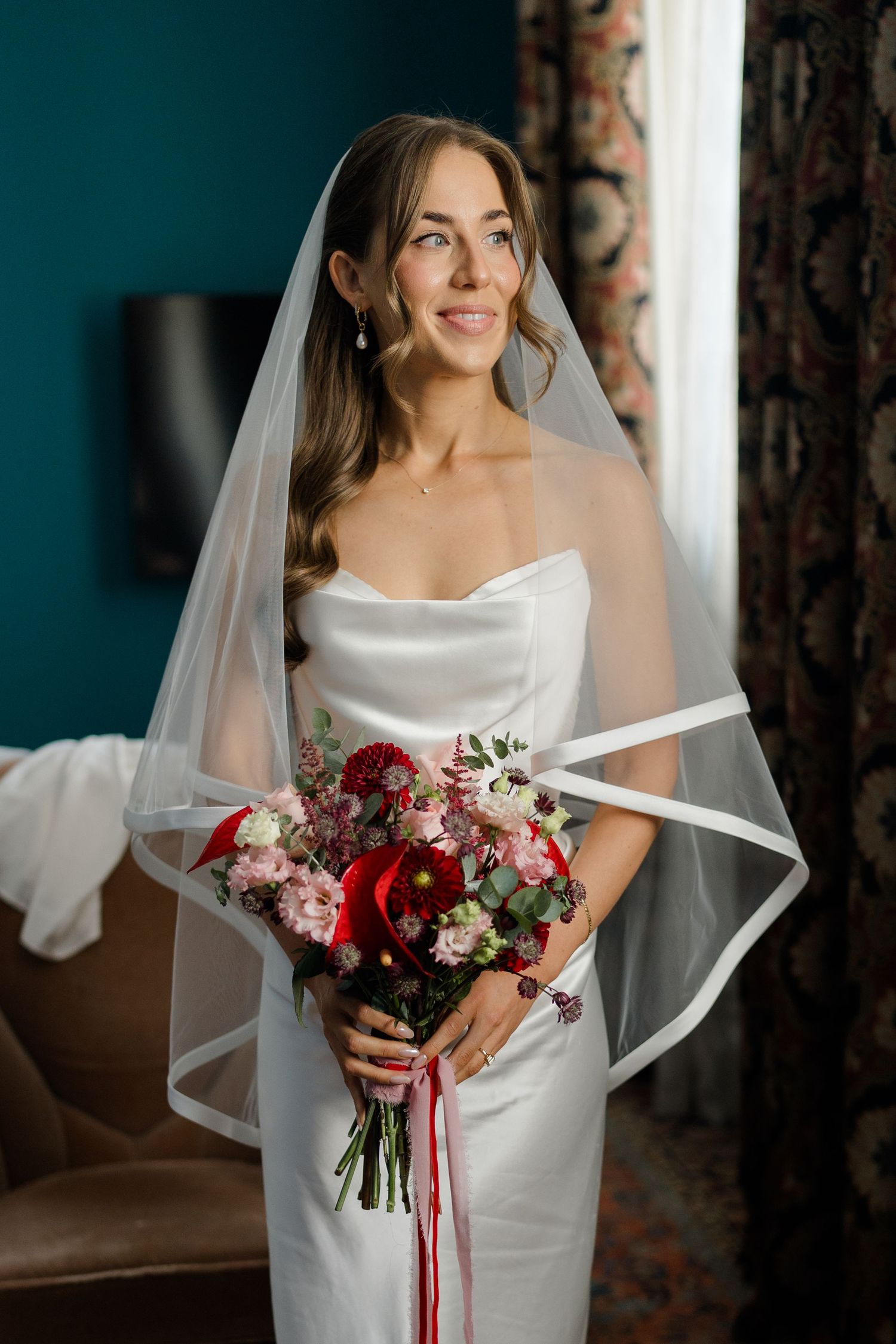 Elegant bride in strapless white gown and veil holding red and pink bouquet, standing by window in bridal suite.