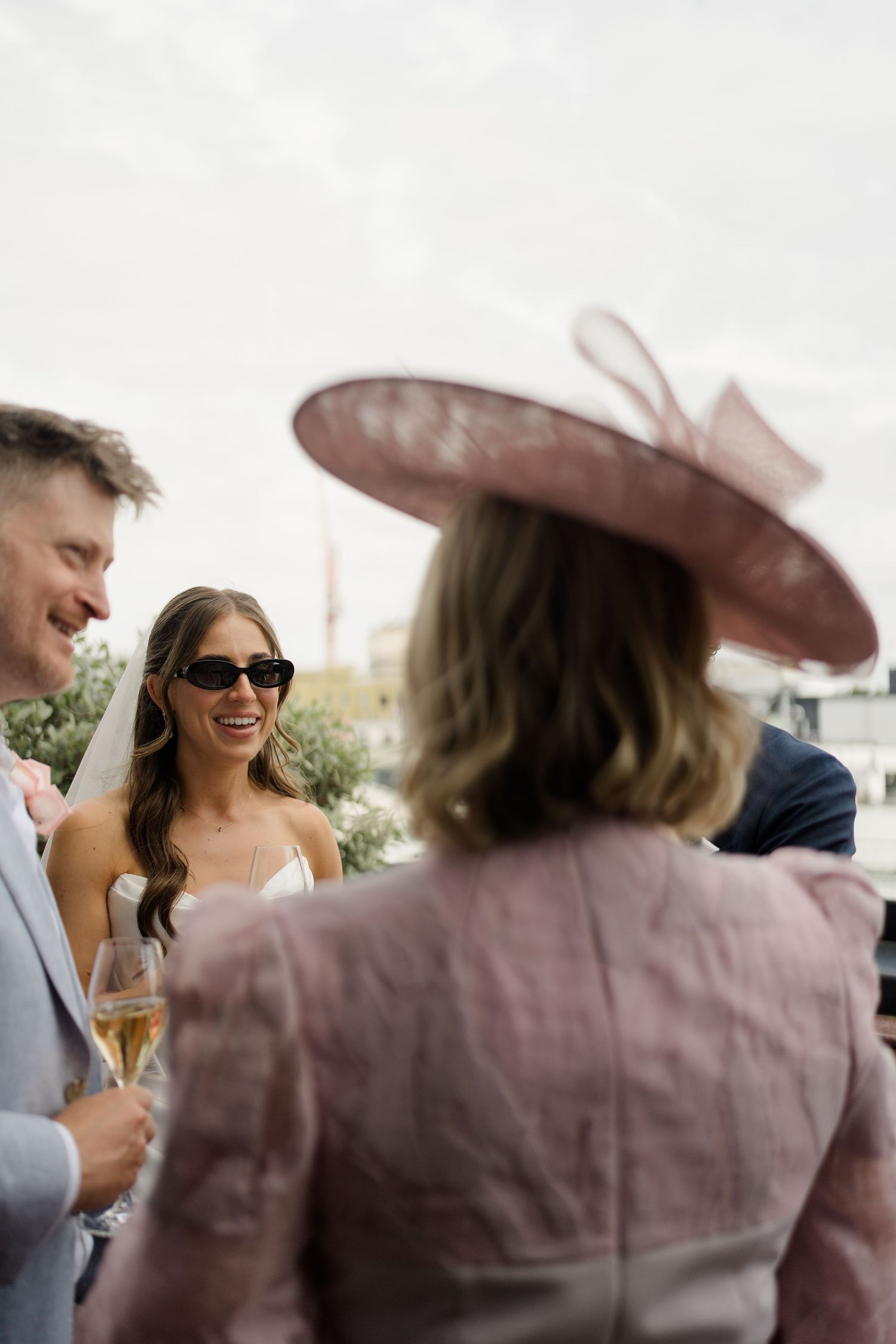 Bride in white dress and sunglasses laughing with wedding guests outdoors, woman in pink fascinator hat in foreground.