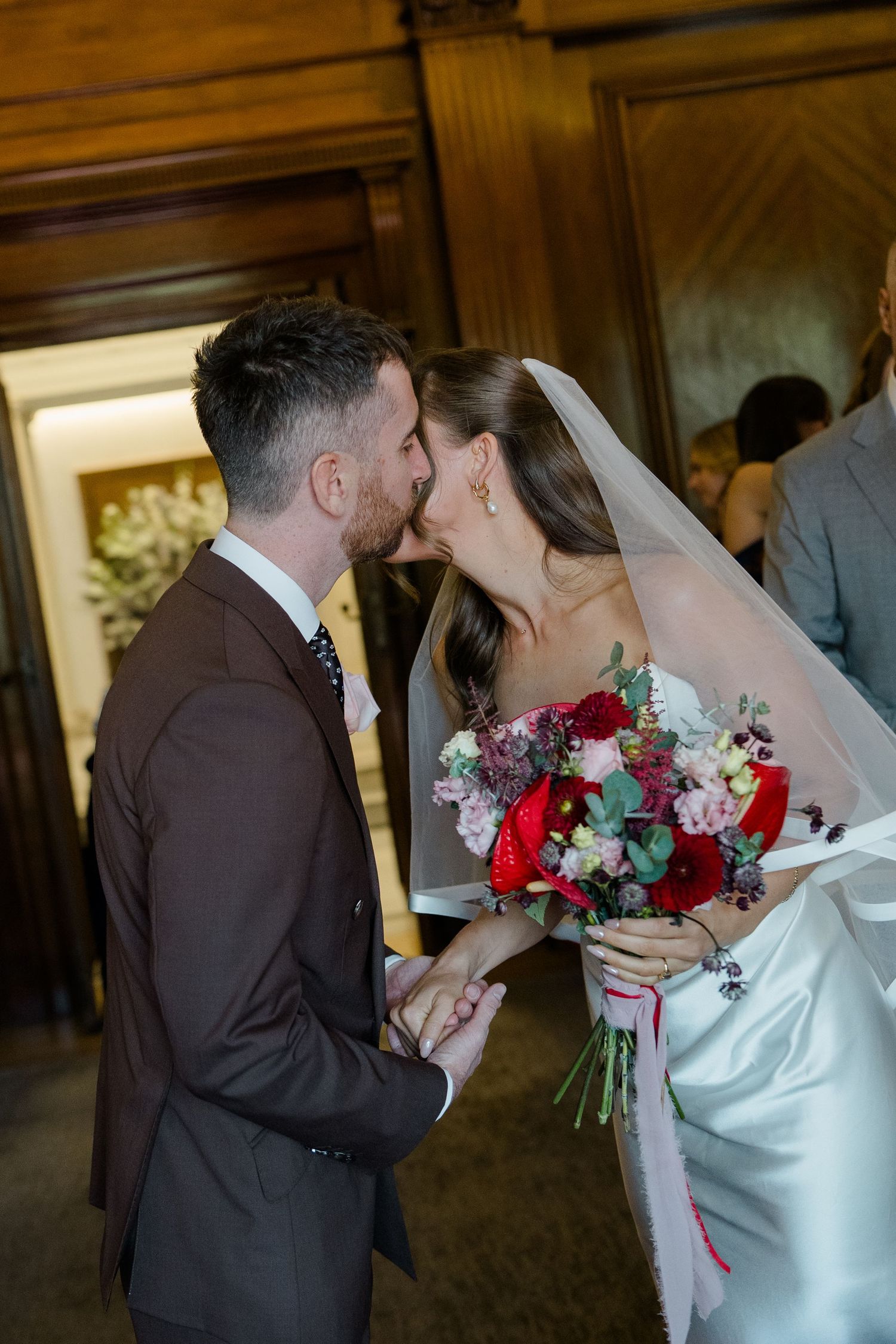 Bride and groom share a tender kiss, bride holding a vibrant red and pink floral bouquet with veil.
