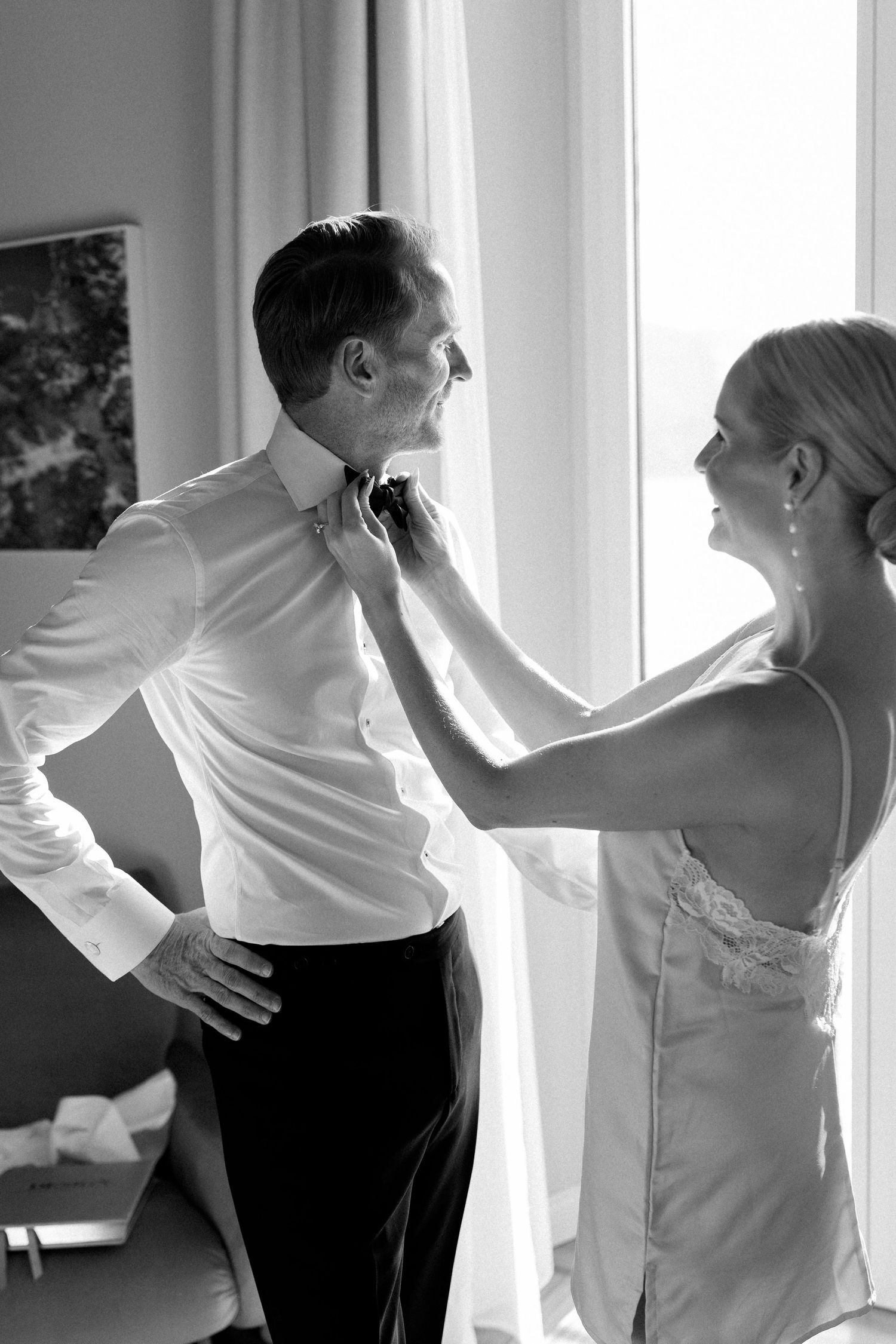 Black and white photo of a smiling bride adjusting her grooms bow tie by a bright window.